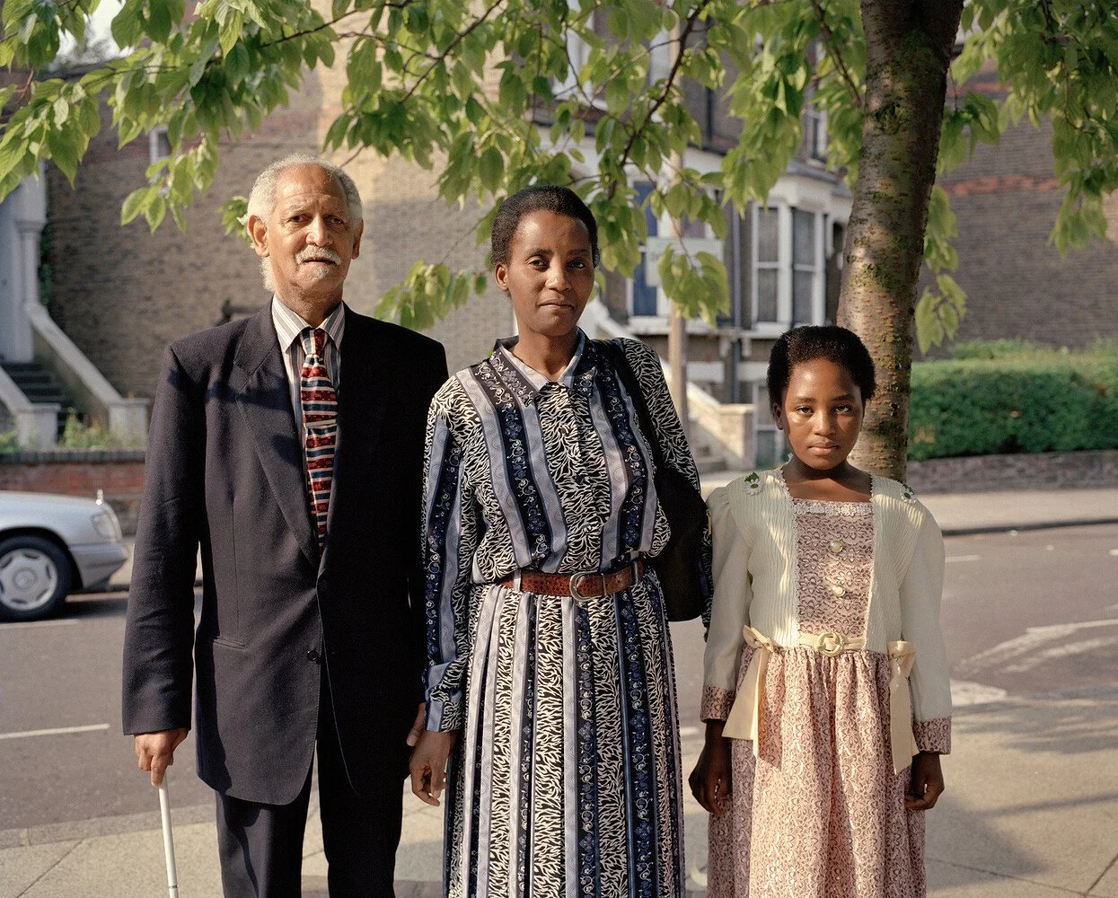Three people standing outdoors in front of a tree and residential houses. An elderly man with a cane on the left, a woman in the middle, and a young girl on the right, dressed in vintage clothing.