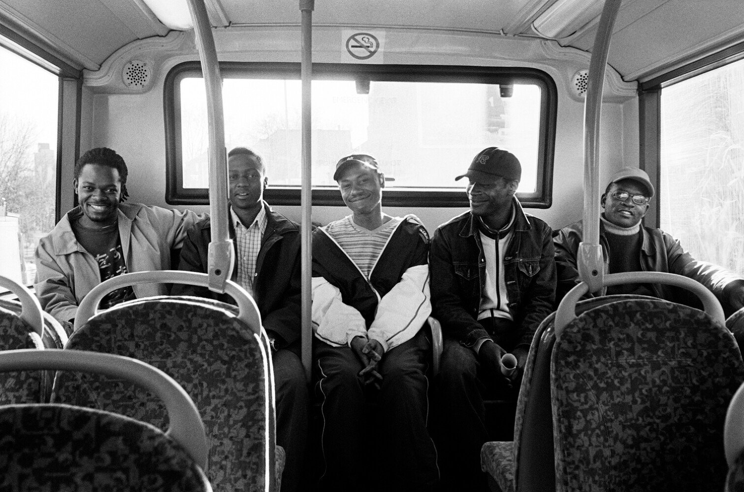 Five men sitting on a bus, smiling at the camera. The bus interior has patterned seats and large windows.