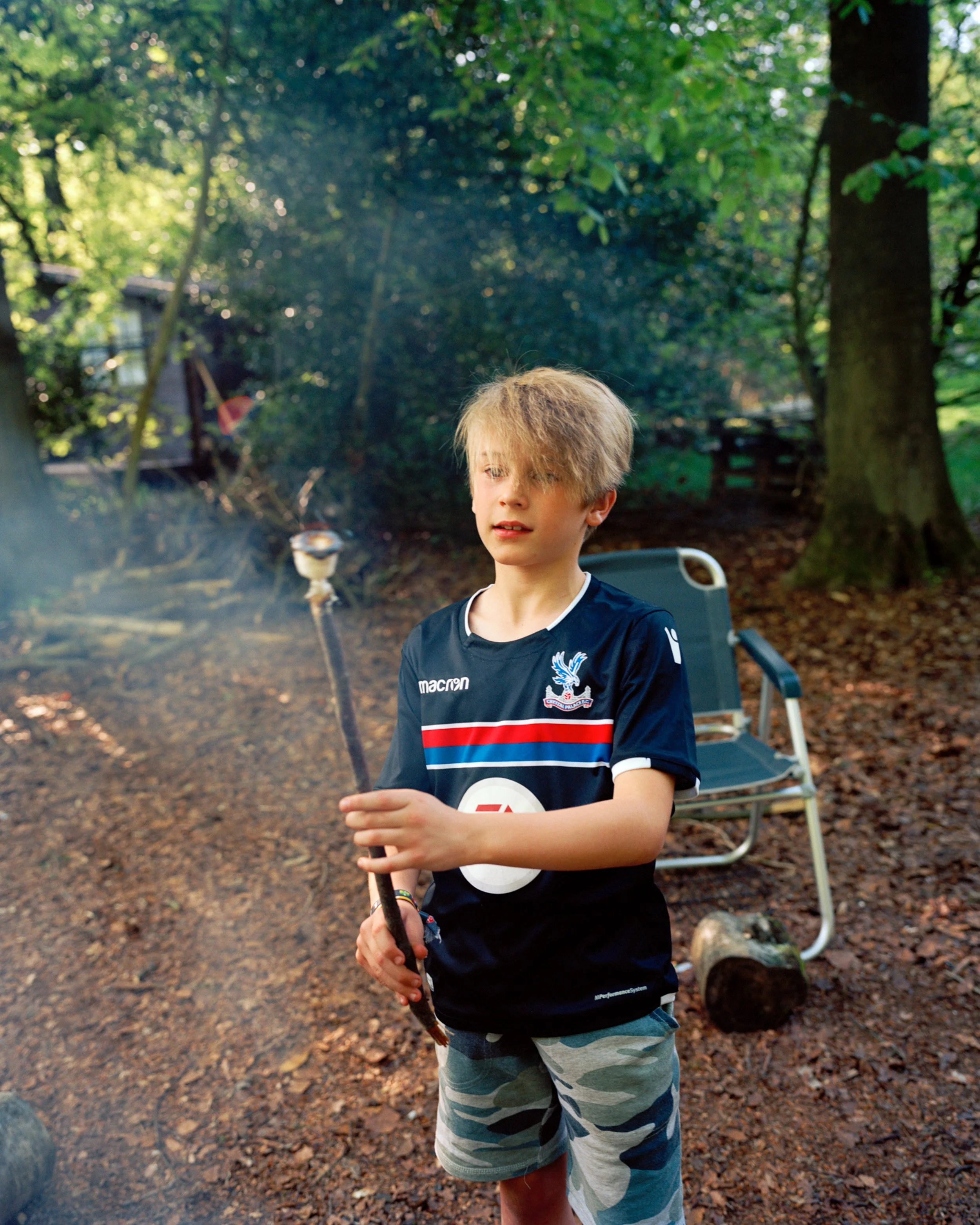 A young boy with messy blonde hair wearing a navy blue sports jersey and camo shorts, holding a stick in a wooded outdoor setting with a chair and logs in the background.