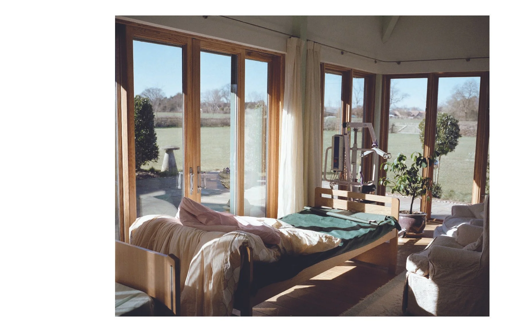 Sunlit hospital room with a bed, medical equipment, and large windows overlooking a grassy outdoor area.