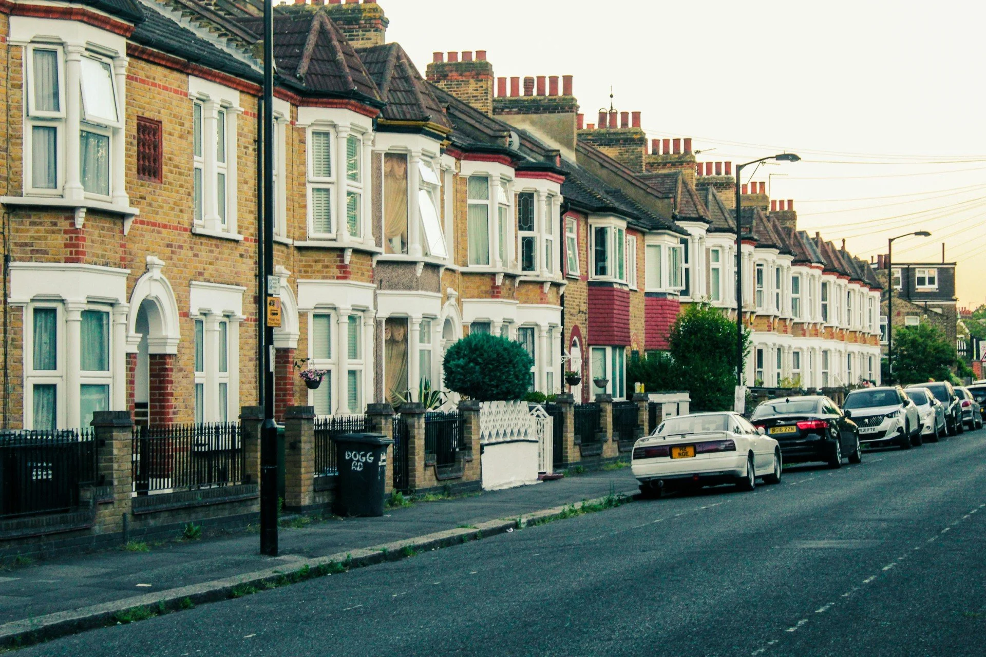 Row of Victorian-style brick houses with white trim and bay windows on a residential street with parked cars and street lamps, during the evening.