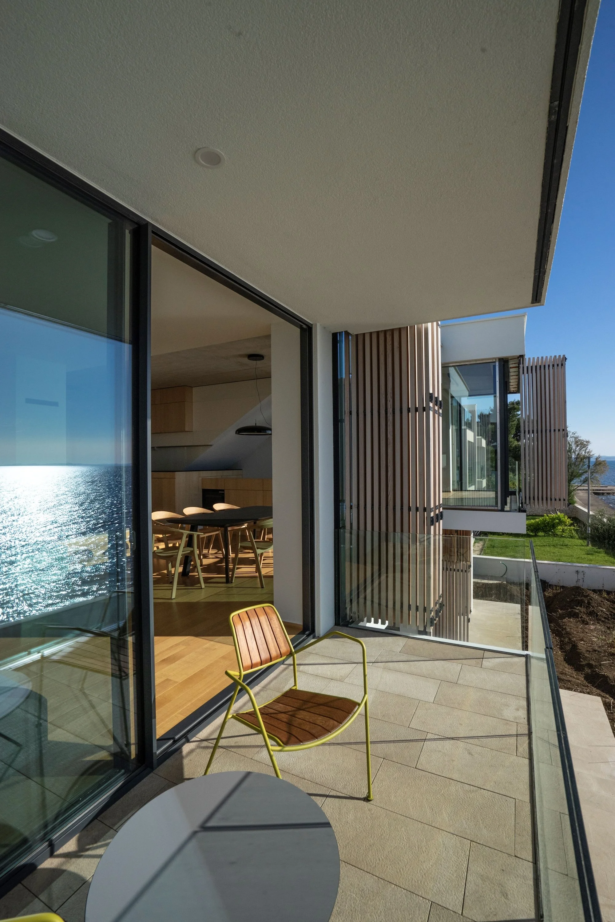 Blick auf die offene Terrasse und das Wohnzimmer eines modernen Hauses mit großen Glasfenstern, Holz- und Metallarchitektur, Blick auf das Wasser im Hintergrund bei sonnigem Wetter.