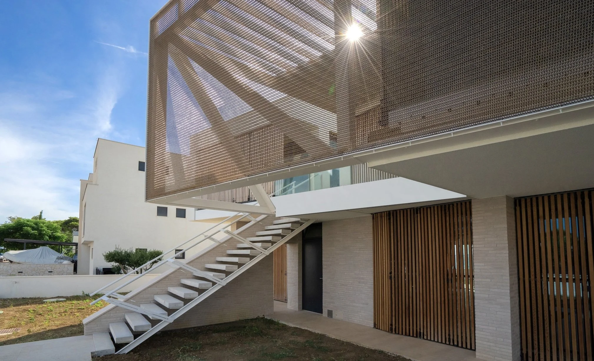 Moderne Gebäudefassade mit weißen Treppen im Vordergrund und einer Holzverkleidung an der Wand. Der Balkon ist mit einer perforierten Metallscheibe geschützt, durch die die Sonne scheint.