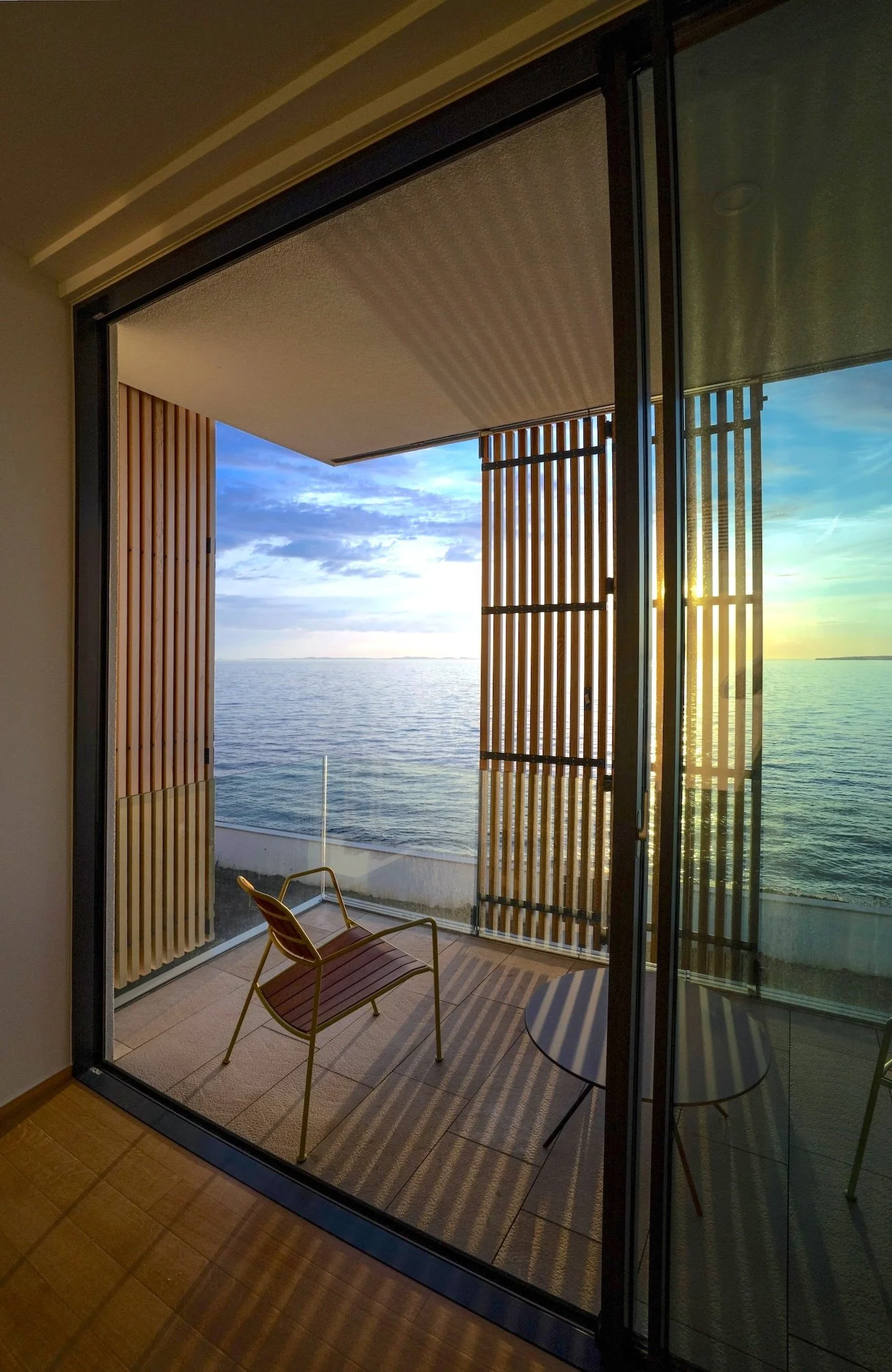 Blick auf eine offene Balkon- oder Terrassentür mit Blick auf den See bei Sonnenuntergang. Es stehen zwei Stühle auf dem Balkon, umgeben von Holzjalousien, die Schatten auf den Boden werfen.