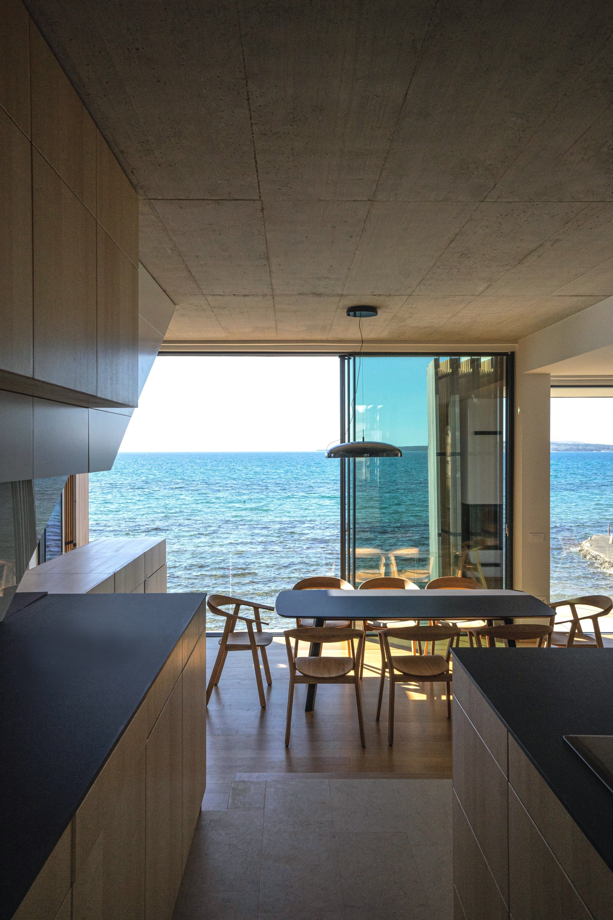 Innenansicht eines modernen Hauses mit Blick auf das Meer durch große Fenster, offener Essbereich mit Holztischen und Stühlen, schlichter Einrichtungsstil.