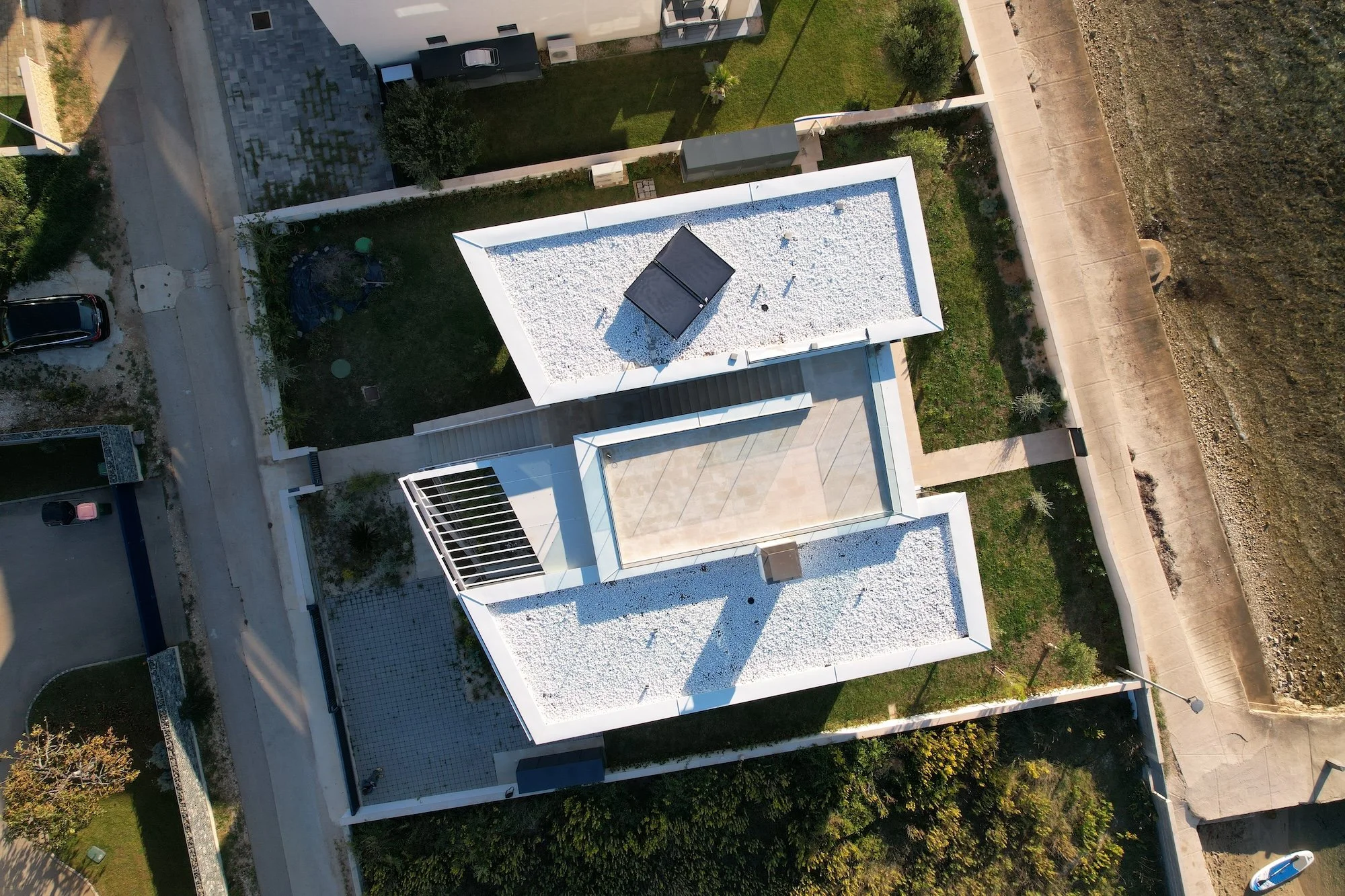 Luftaufnahme eines modernen weißen Hauses mit Flachdach, das mit weißen Steinen bedeckt ist. Das Haus hat einen Balkon und ist von Gärten, Zäunen und Straßen umgeben.