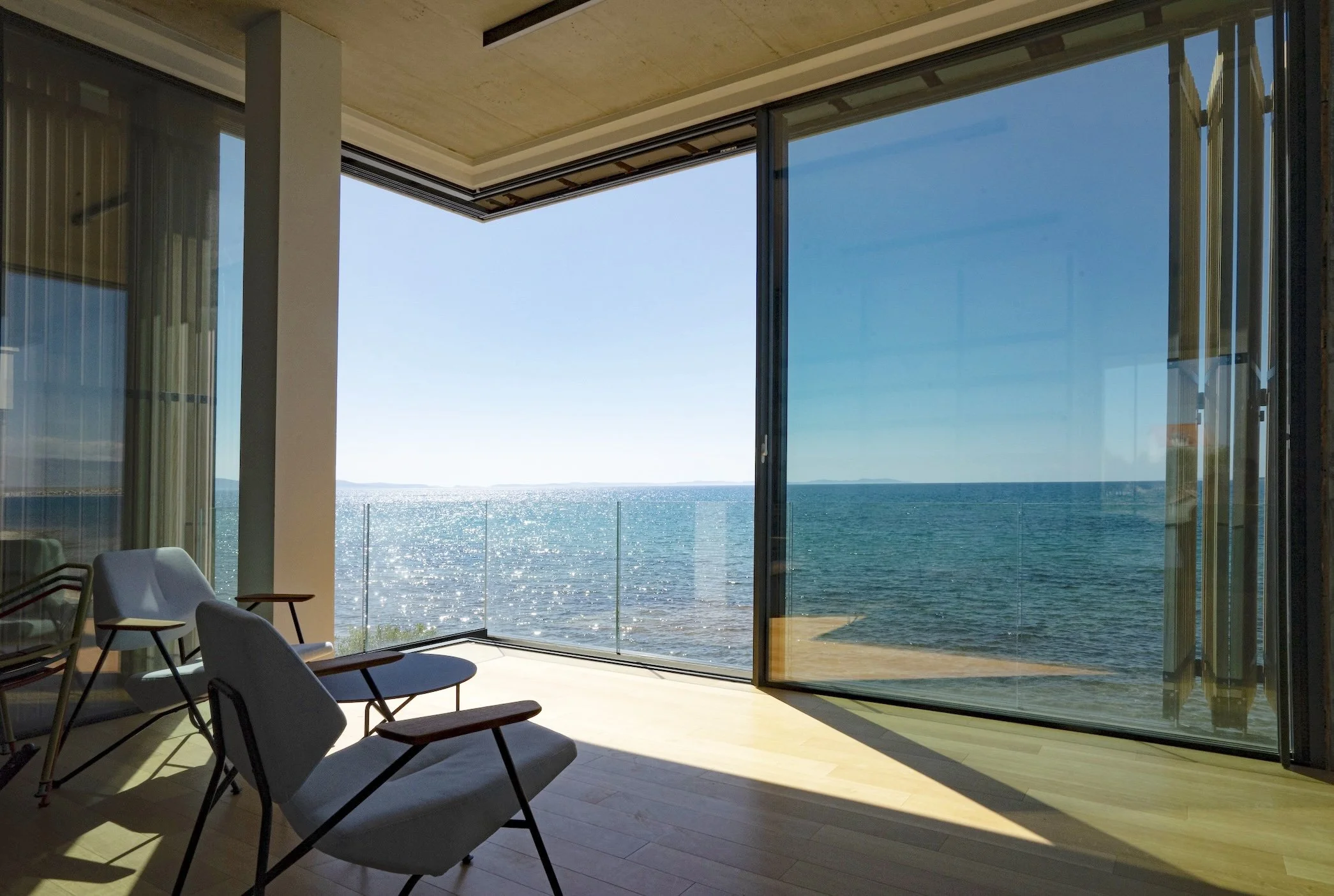 Moderne Raum mit großen Glasfenstern, Blick aufs Meer, einige Stühle im Vordergrund.