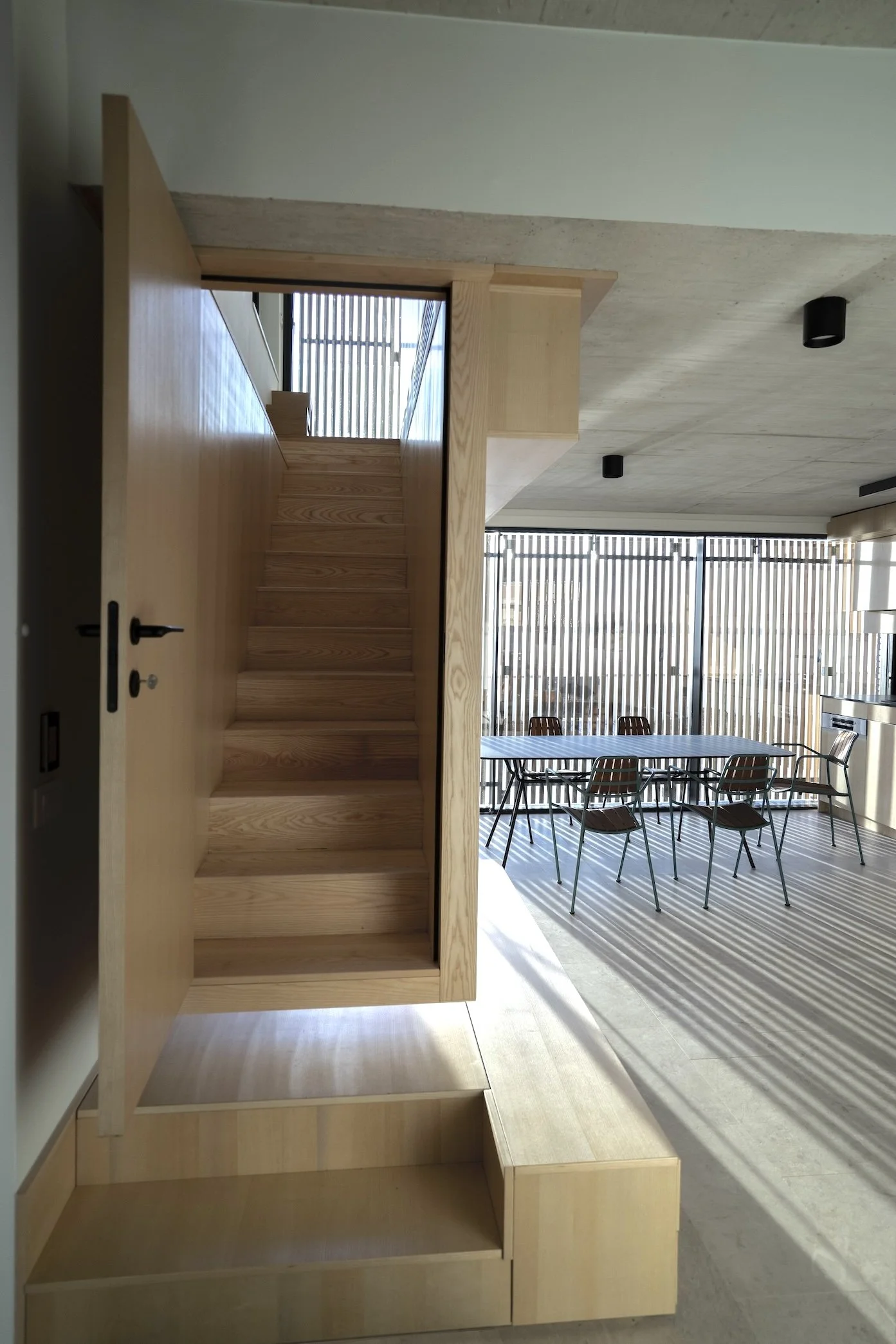 Innenraum mit Holztreppe, gläsernen Wänden, Essbereich mit Tischen und Stühlen, Sonne scheint durch Fenster, moderner minimalistischer Stil.