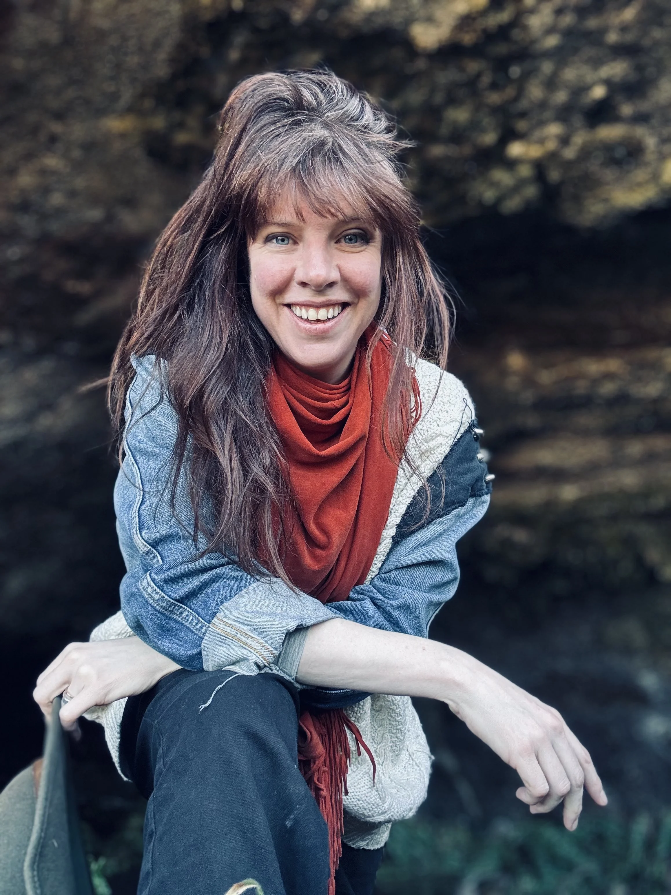 A woman with long brown hair and blue eyes smiling outdoors, wearing a denim jacket, red scarf, and black pants, with a blurred natural background.