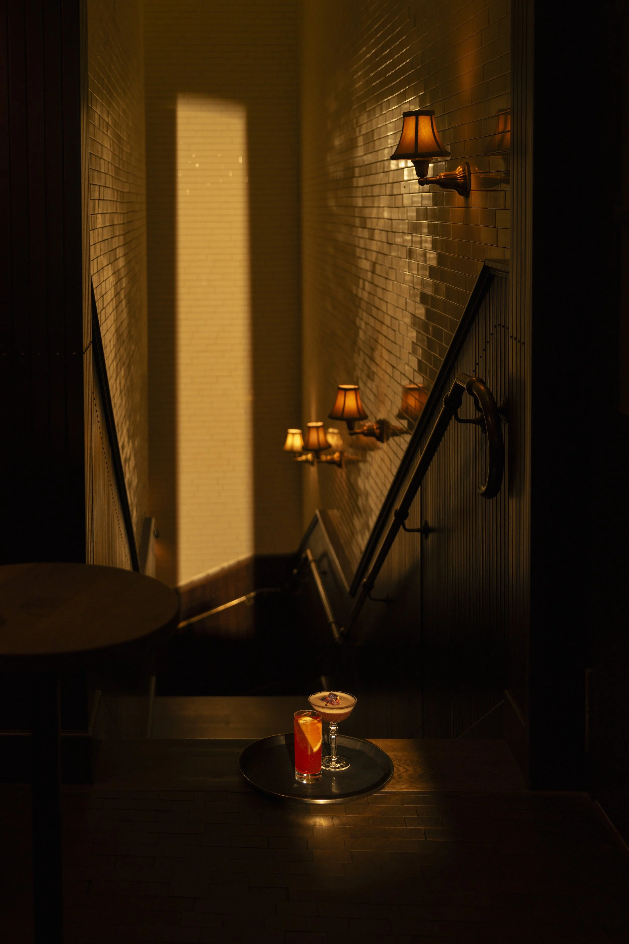 A dimly lit staircase with wall-mounted lamps, and a table in foreground with two cocktails.