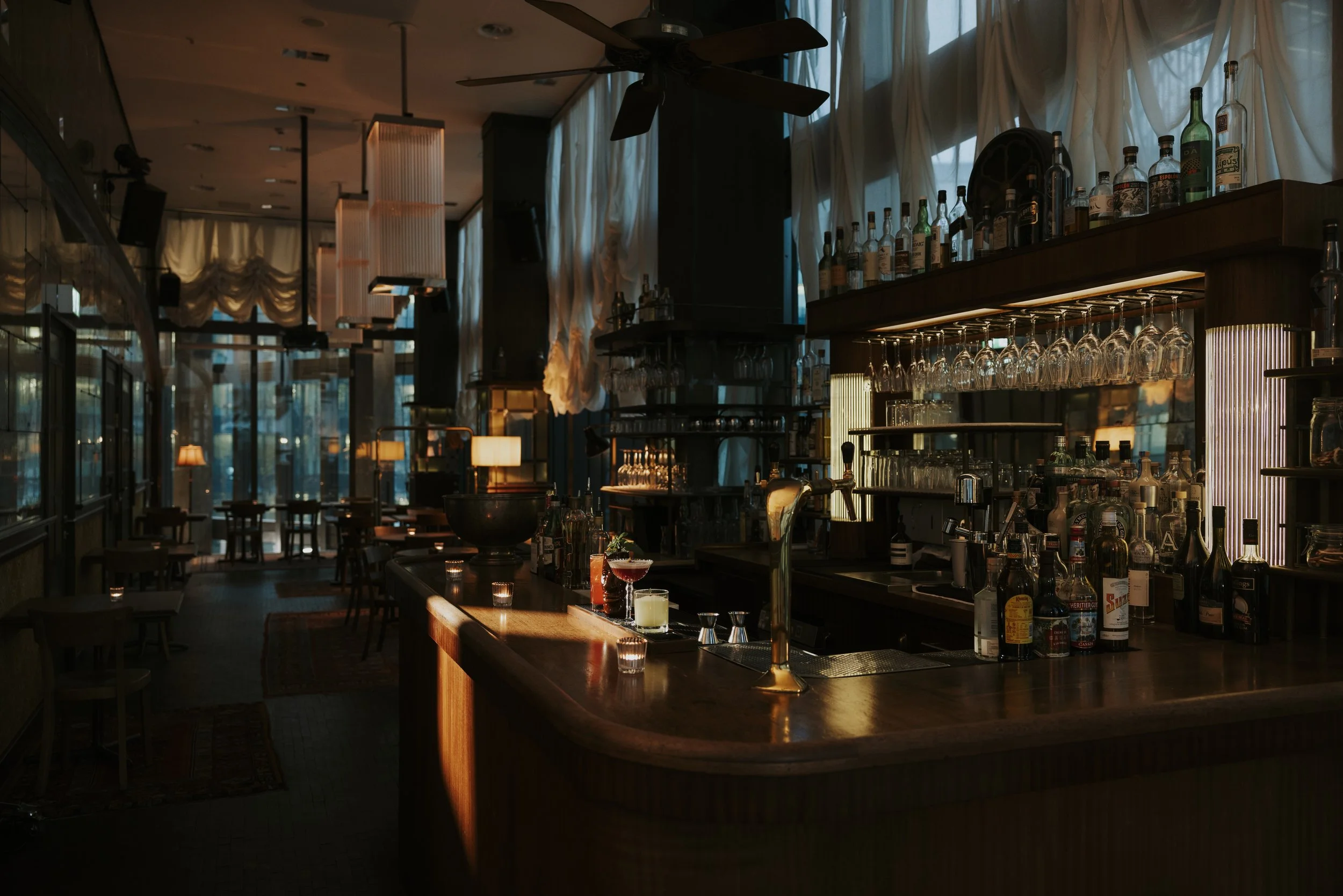 Empty bar with dim lighting, liquor bottles on shelves, glasses hanging upside down, illuminated by soft light, with tables and chairs in the background.