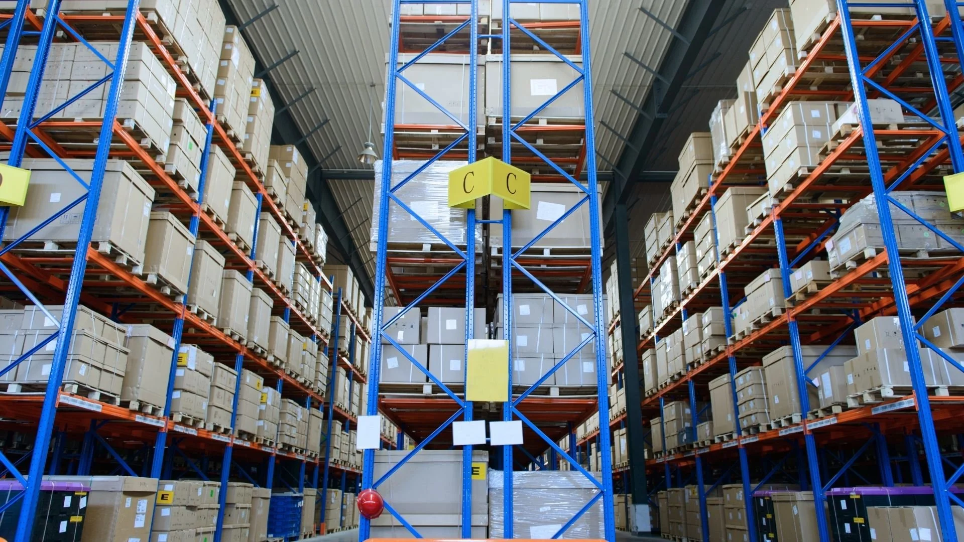 View of a large warehouse with high metal shelving filled with stacked cardboard boxes, some wrapped in plastic, organized on multiple levels.