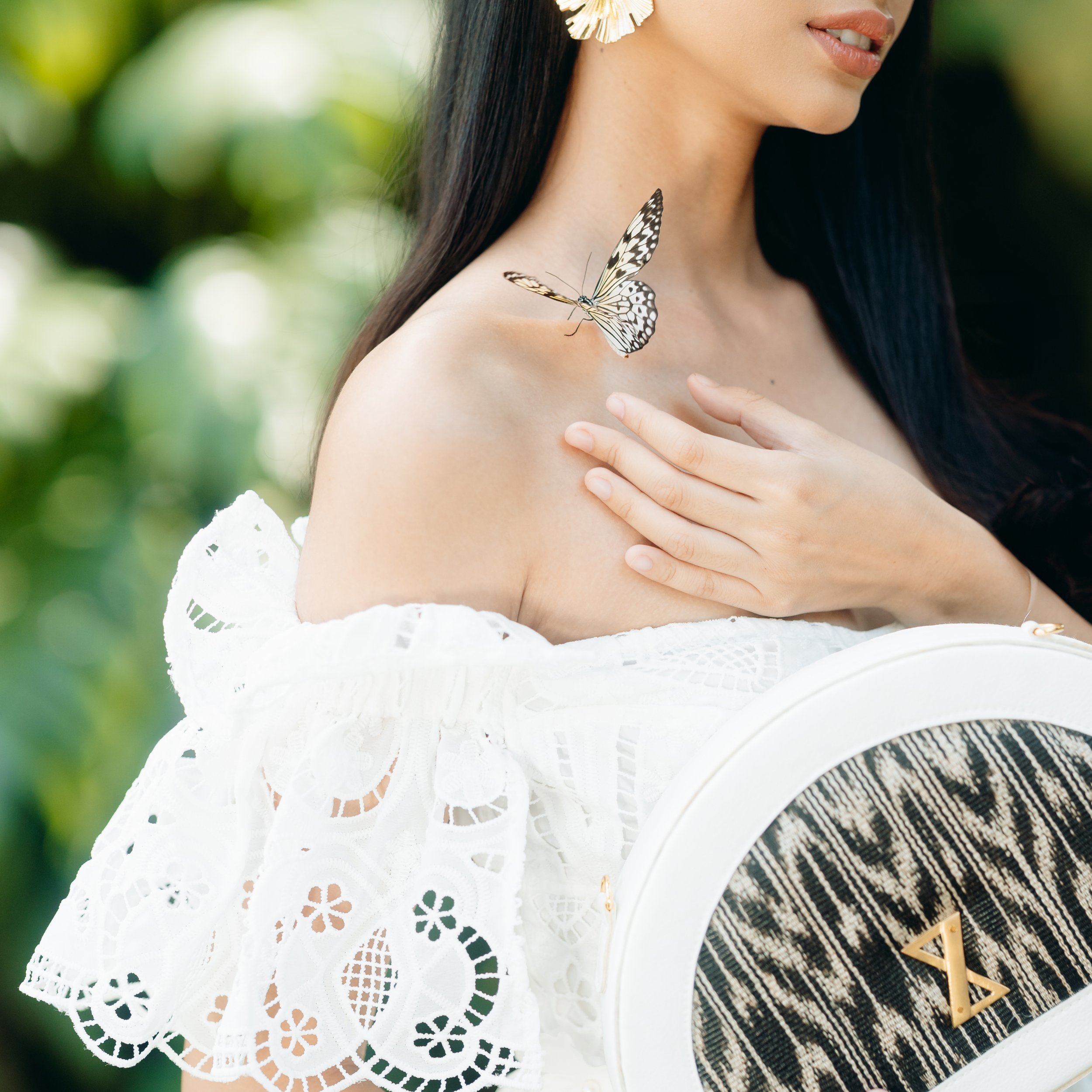 A woman in white lace off-the-shoulder dress outdoors, touching her shoulder as a butterfly lands nearby.