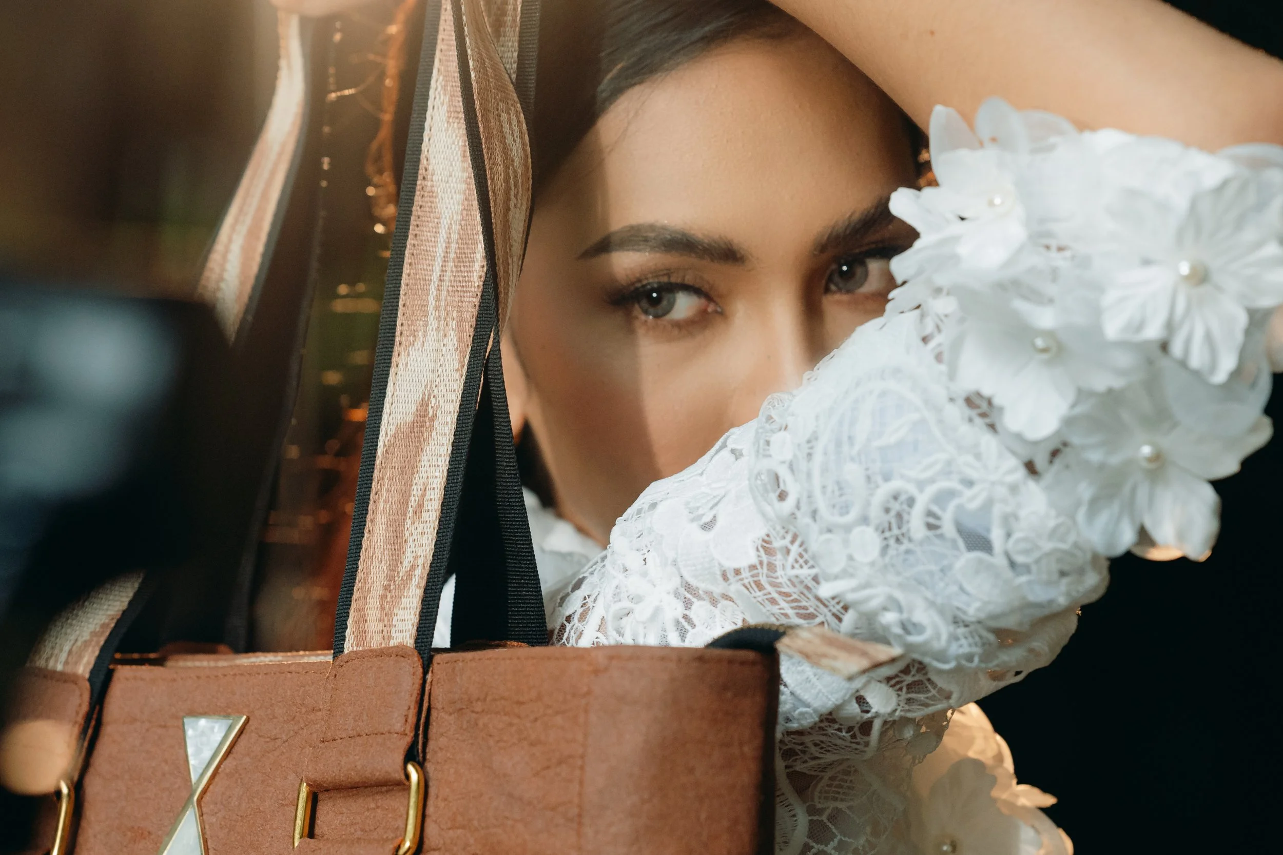 A woman with light eyes and dark hair, partially obscured by a cream-colored lace sleeve with floral accents and pearl details, peering out from behind a brown handbag with striped handles.