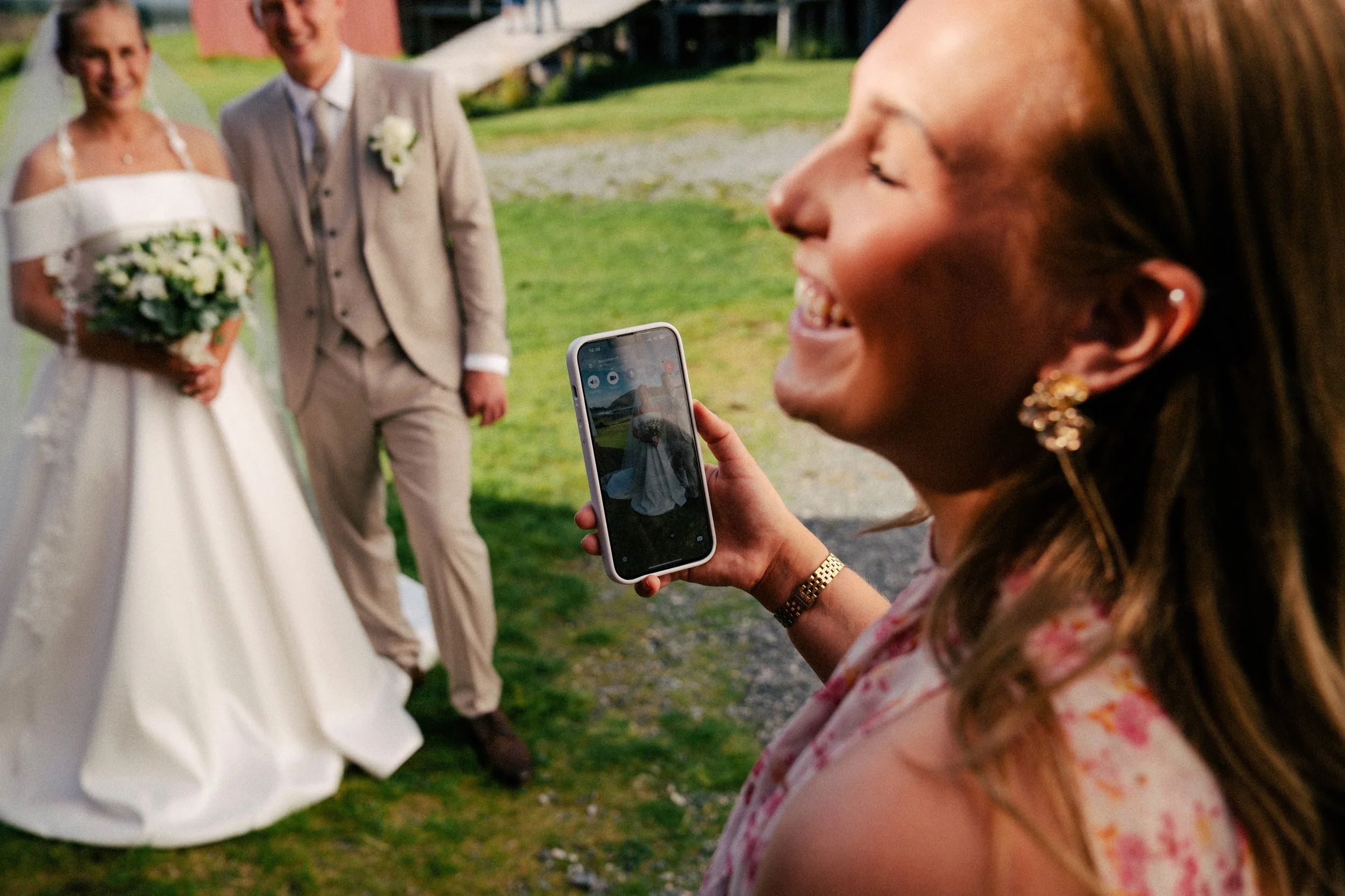 En kvinne tar et bilde av et brudepar med telefonen sin, mens hun smiler og ler.