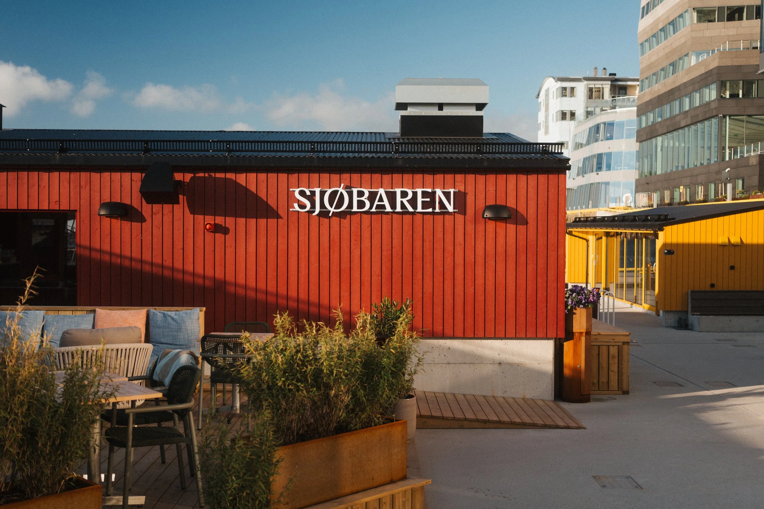 Et utendørs område med en rødfarget bygning med skiltet 'Sjøbaren', restaurant- eller barfasiliteter, og bord med stoler foran. Moderne bygninger i bakgrunnen under en blå himmel.