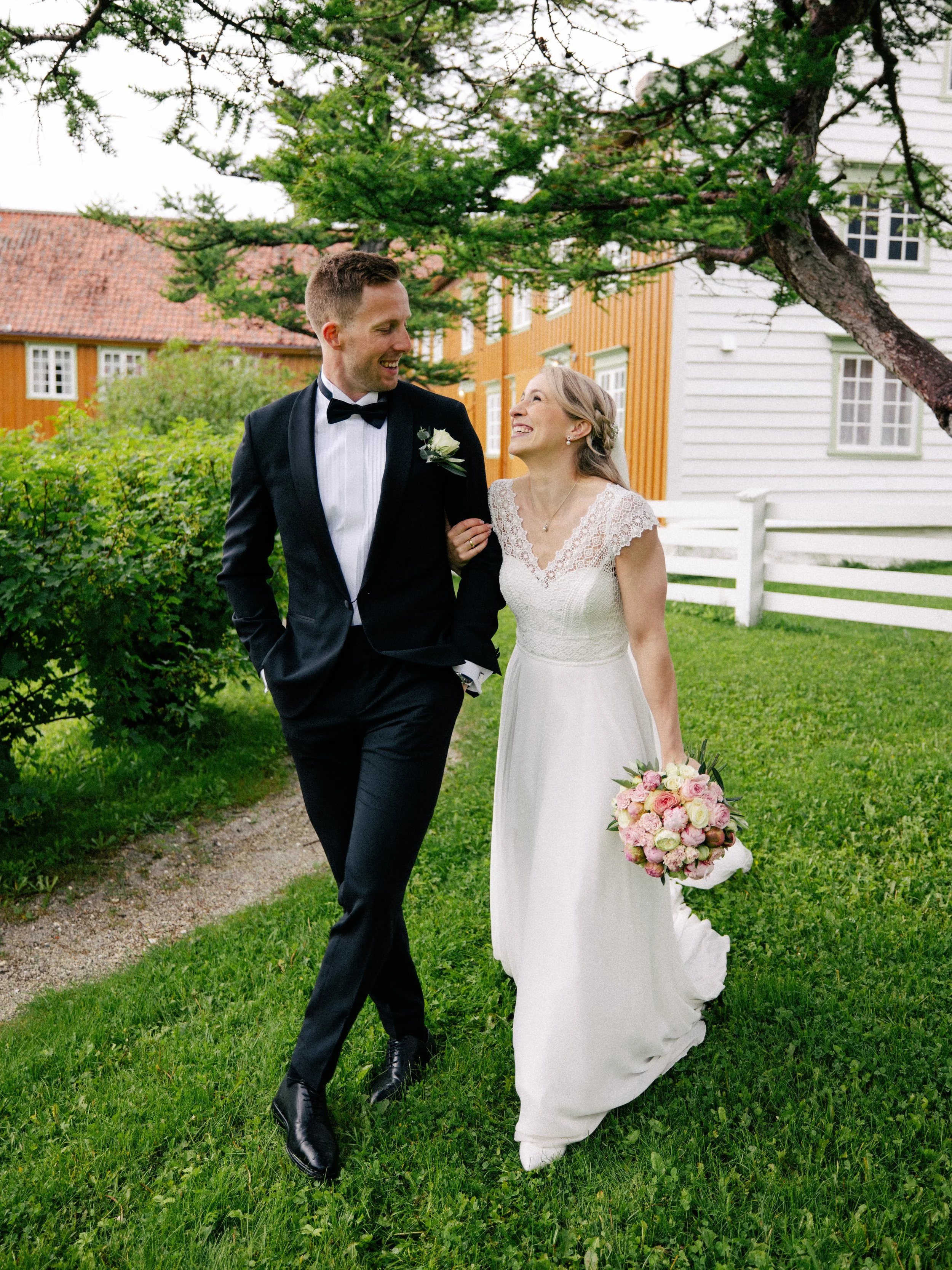 Et par i brudekjole og smoking går hånd i hånd på en grønn gresslette med trær og hus i bakgrunnen.