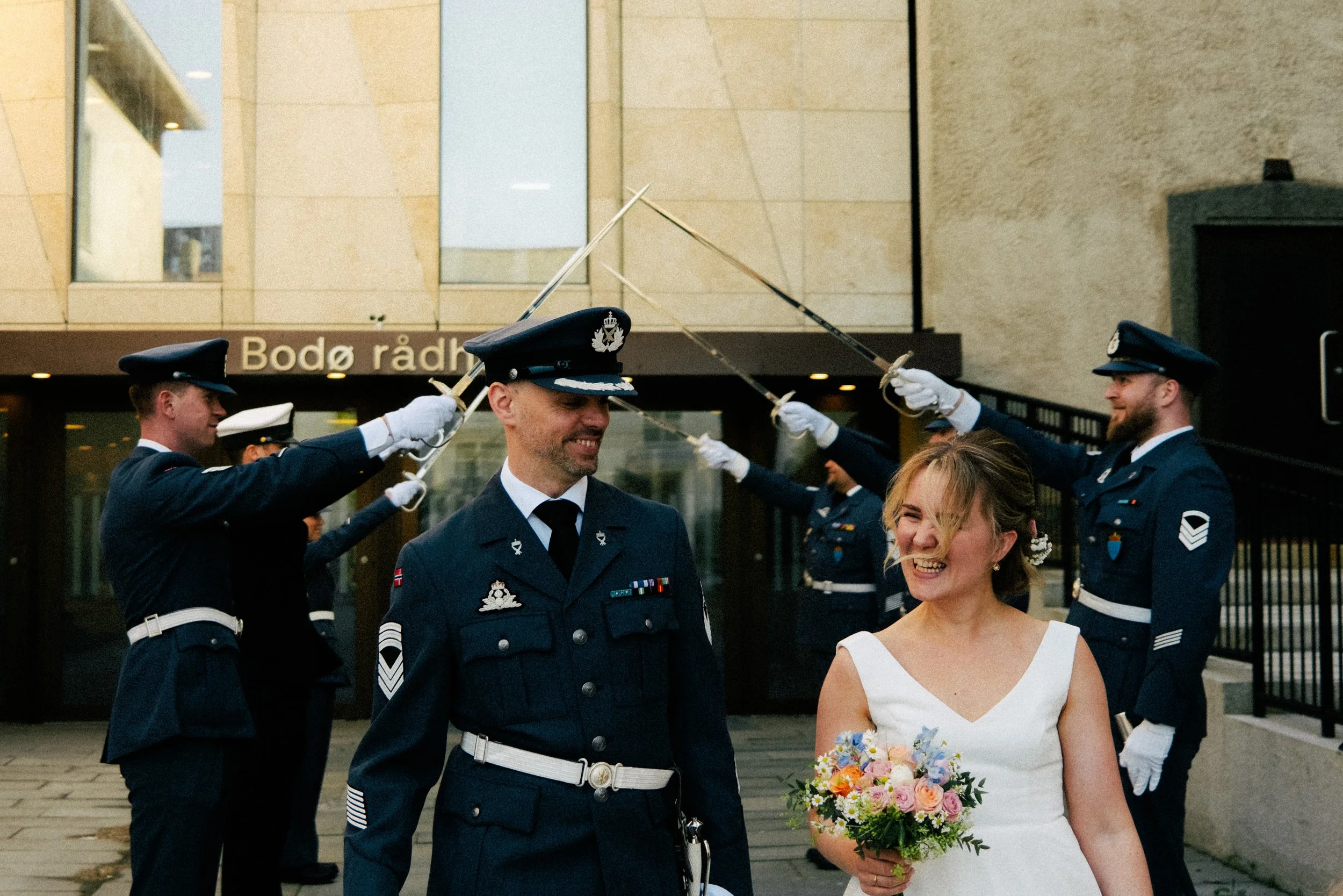 Ung kvinne i en hvit kjole holder en blomsterbukett og smiler mens en mann i militæruniform står foran henne. Flere menn i militæruniformer løfter sabler i en seremoni foran bygningen Bodø rådhus.