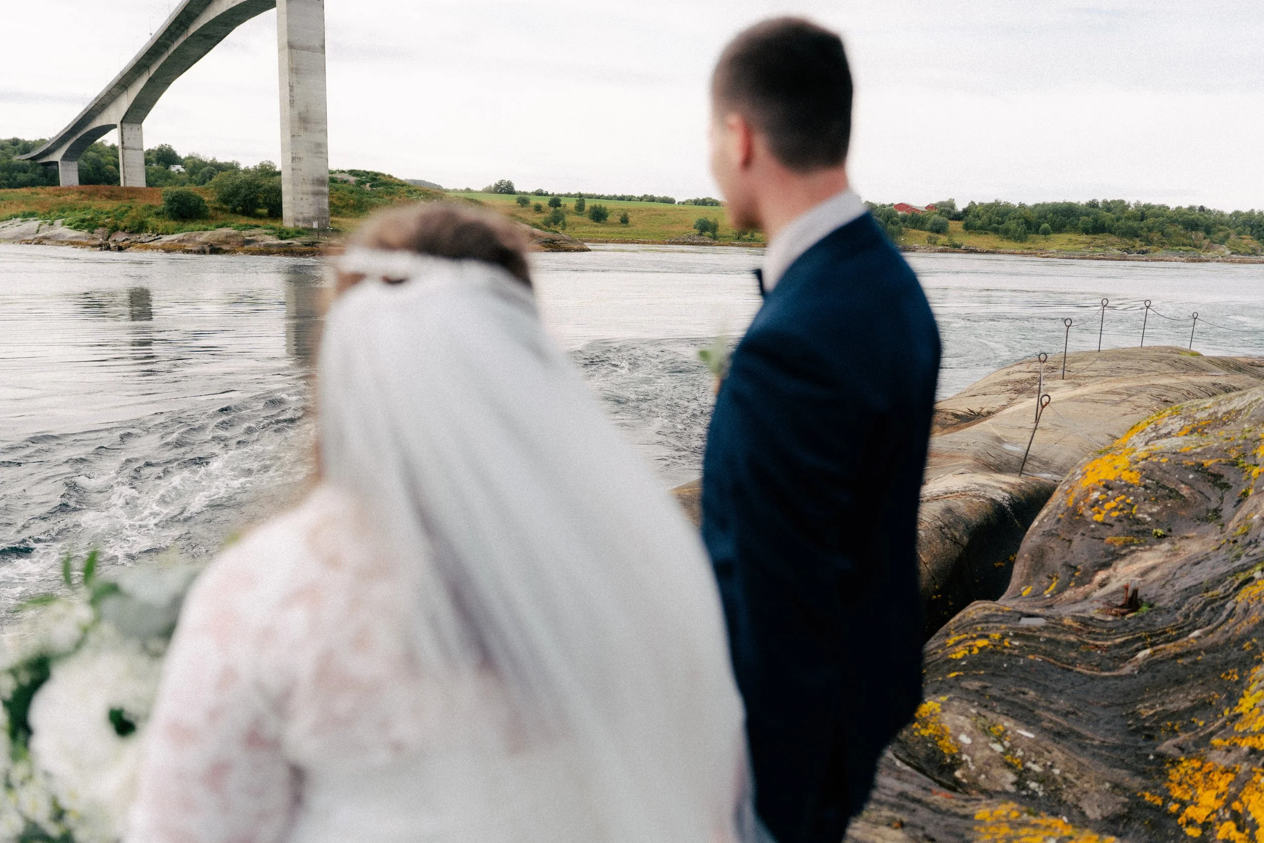To personer står ved en sjø med en bro i bakgrunnen. Den ene personen har på seg en hvit dress og er omgitt av blomster, den andre har på seg en mørk dress.