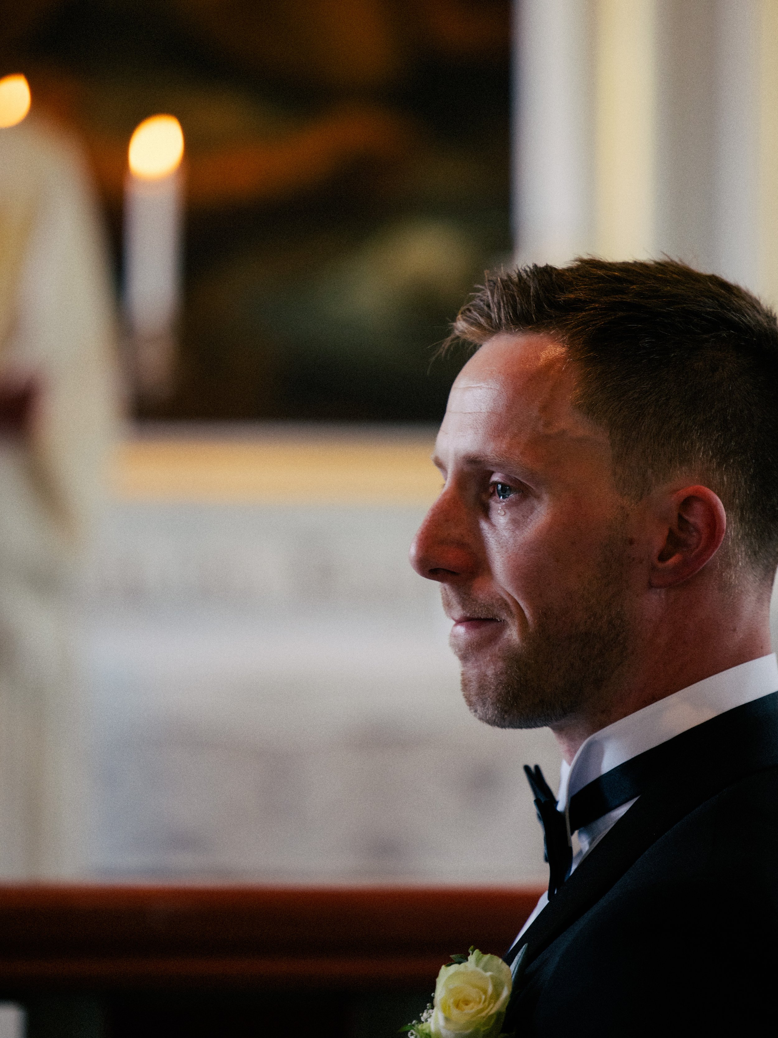 Nærbilde av en mann i sort dress med hvit skjorte og svart sløyfe, med en hvit rosenboutonniere på brystlommen.