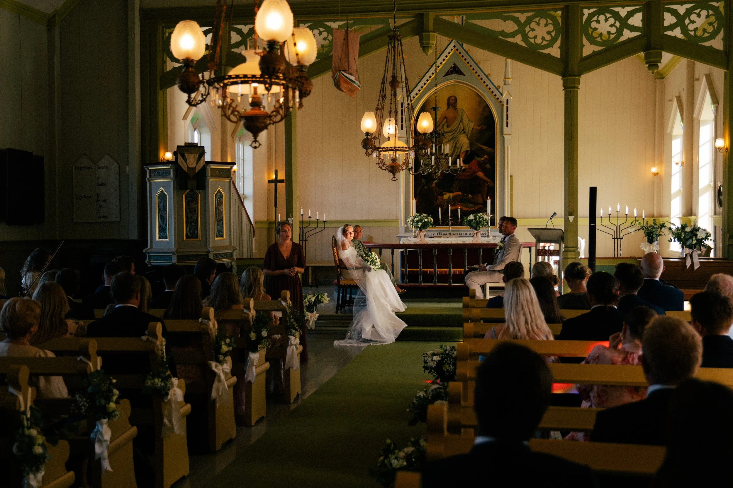 Et bryllup i en kirke med bruden og brudgommen sittende foran alteret, omgitt av familie og venner. Typiske kirkeelementer som lysestaker, kors og religiøse bilder er synlige.