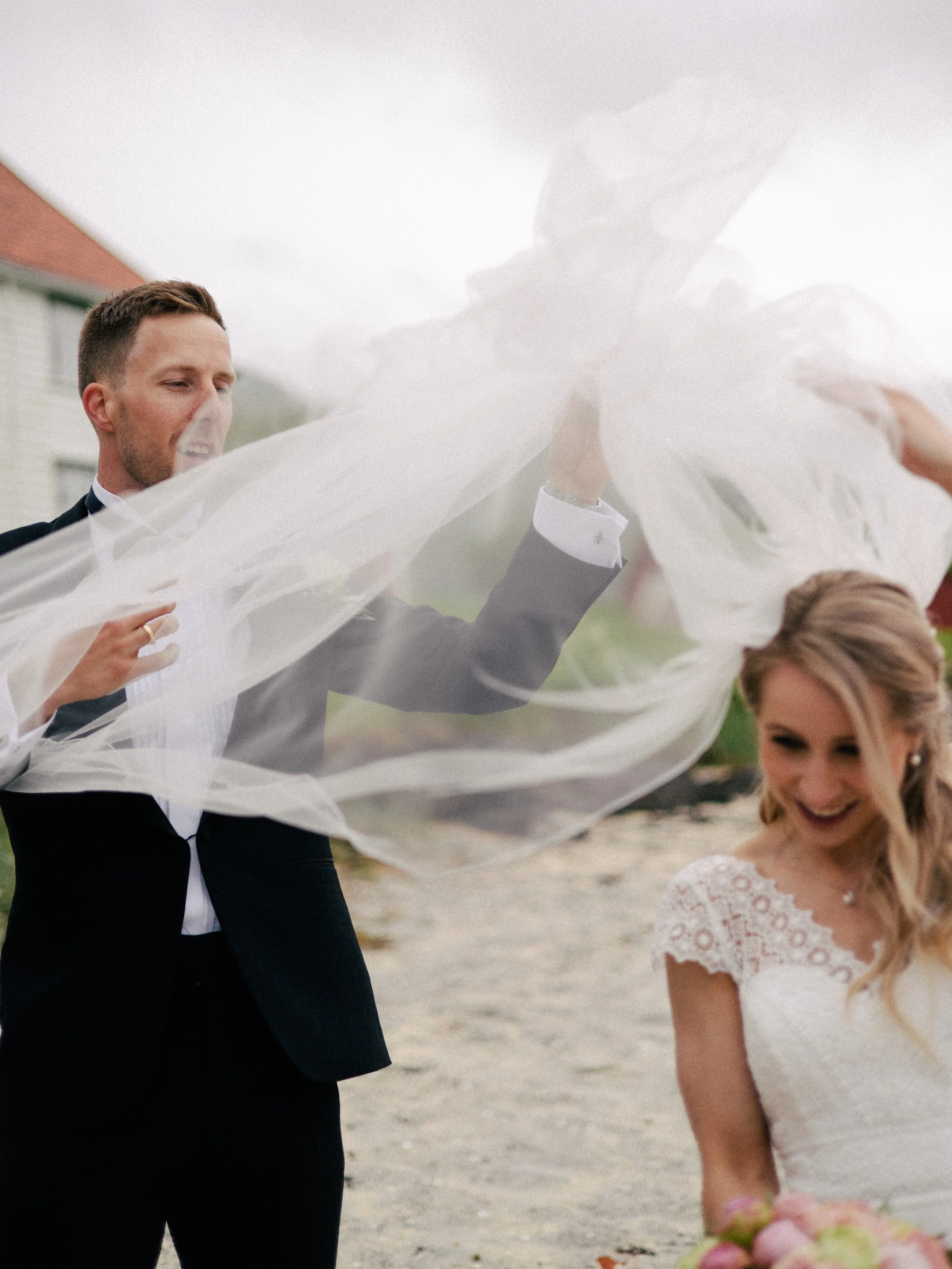 Bildet viser en mann i formell svart dress og en kvinne i en hvit brudekjole som smiler og ler på stranden. Mannen holder en hvit brudekjole eller et stykke av stoff, og kvinnen er i ferd med å plukke opp en bukett med blomster. Det er en trang og gl