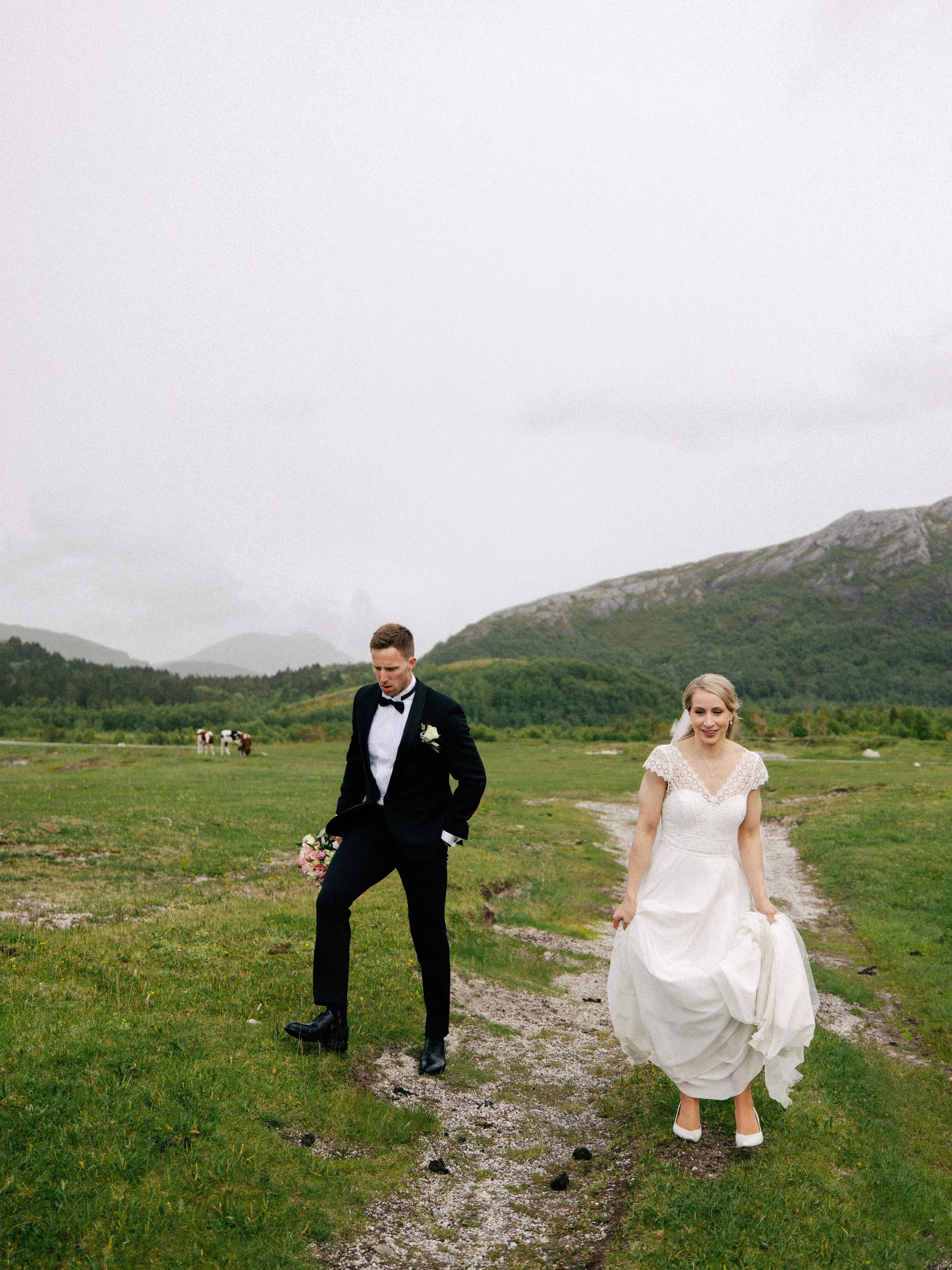 En brud og en brudgom går på en grusvei i et grønt landskap på Kjerringøy utenfor Bodø. Bruden har på seg kjole og høye hæler, og brudgommen har på seg en svart dress og slips. I bakgrunnen kan man se noen kyr på en eng og fjell.