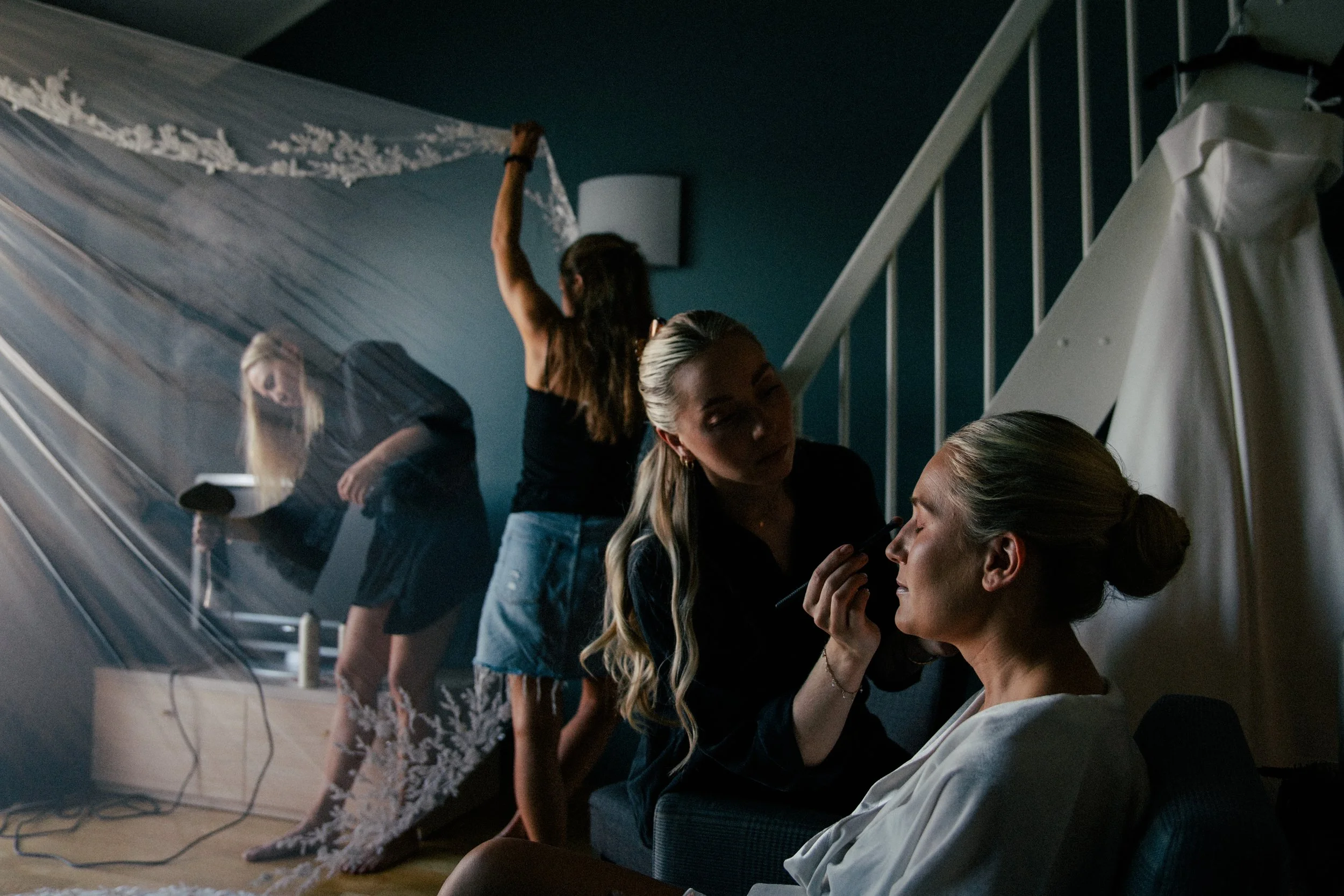 En gruppe kvinner som forbereder seg til en bryllupsseremoni, med noen som hjelper bruden med påkledning og sminke, i et rom med trapp og dekorative elementer.