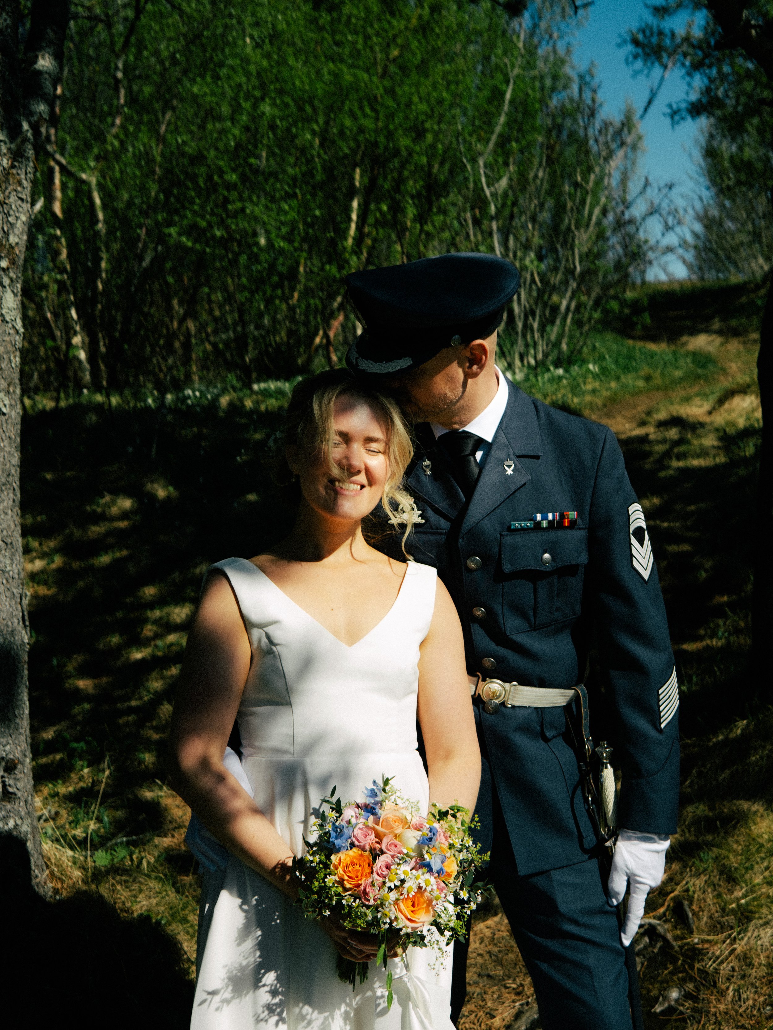En kvinne i brudekjole holder en blomsterbukett, mens en mann i militæruniform kysser hennes panne i en skog. Været er klart, og det er grønne trær rundt dem.