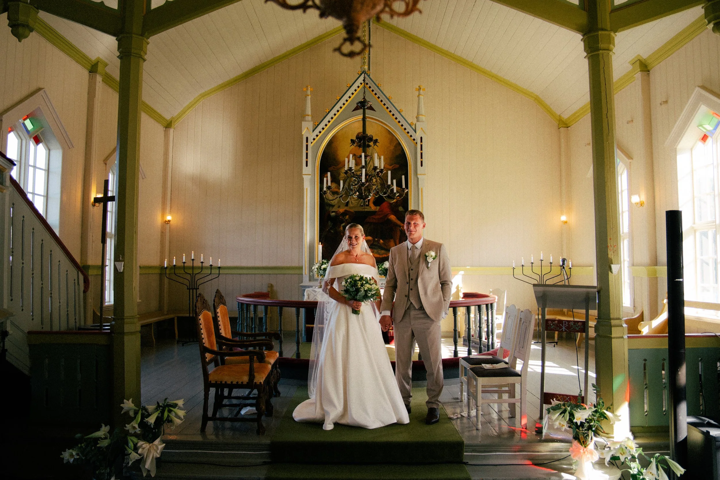 En brud og en brudgom står sammen inne i en kirke, feirer sitt bryllup. De smiler og holder hender. Det er vakker dekorasjon med blomster rundt dem, og kirken er innredet med tradisjonelle elementer som lysekroner og malerier.