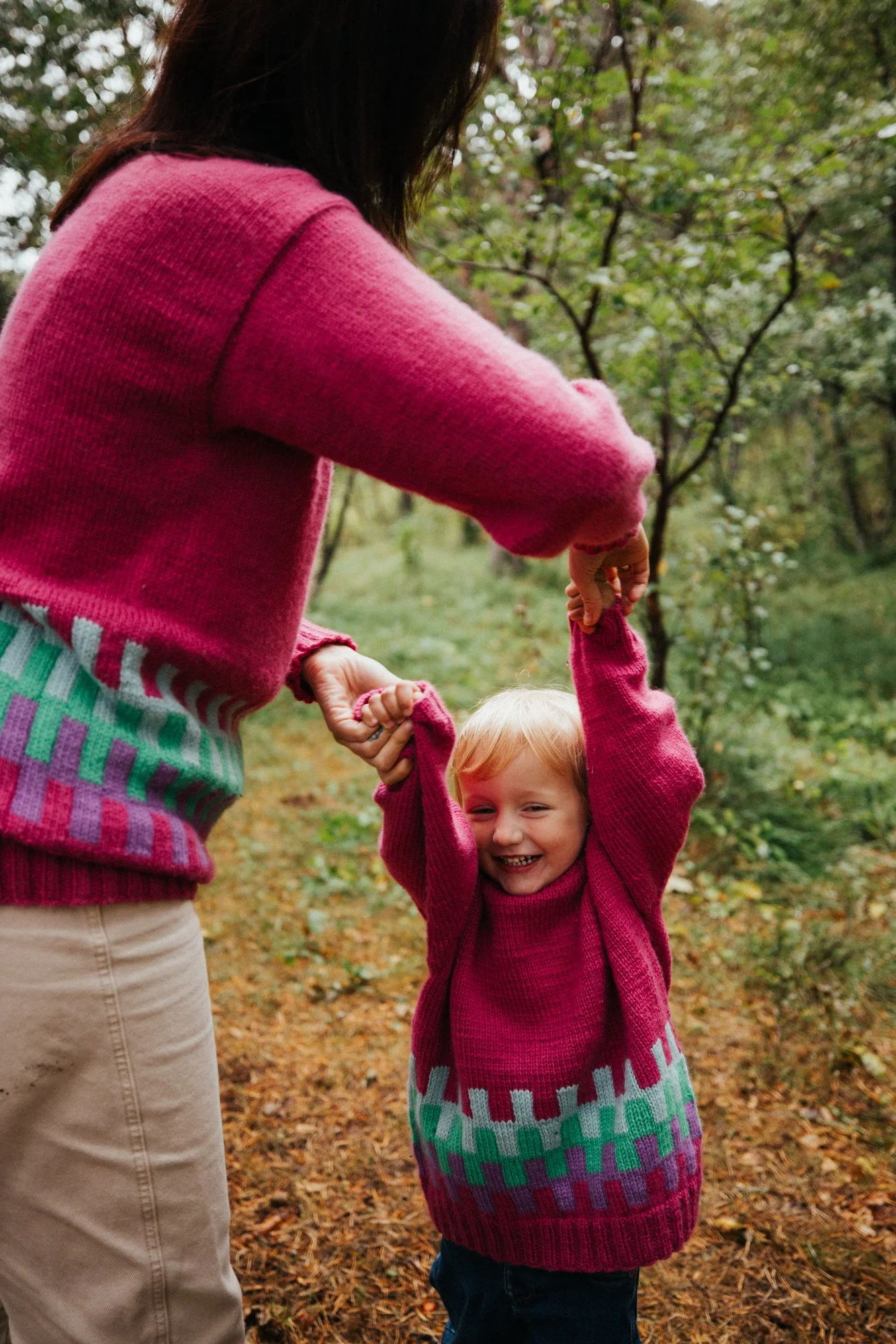 A woman and a young girl playing together outdoors in a forest. The woman is holding the girl's hands as she spins around, both are smiling and dressed in pink sweaters.