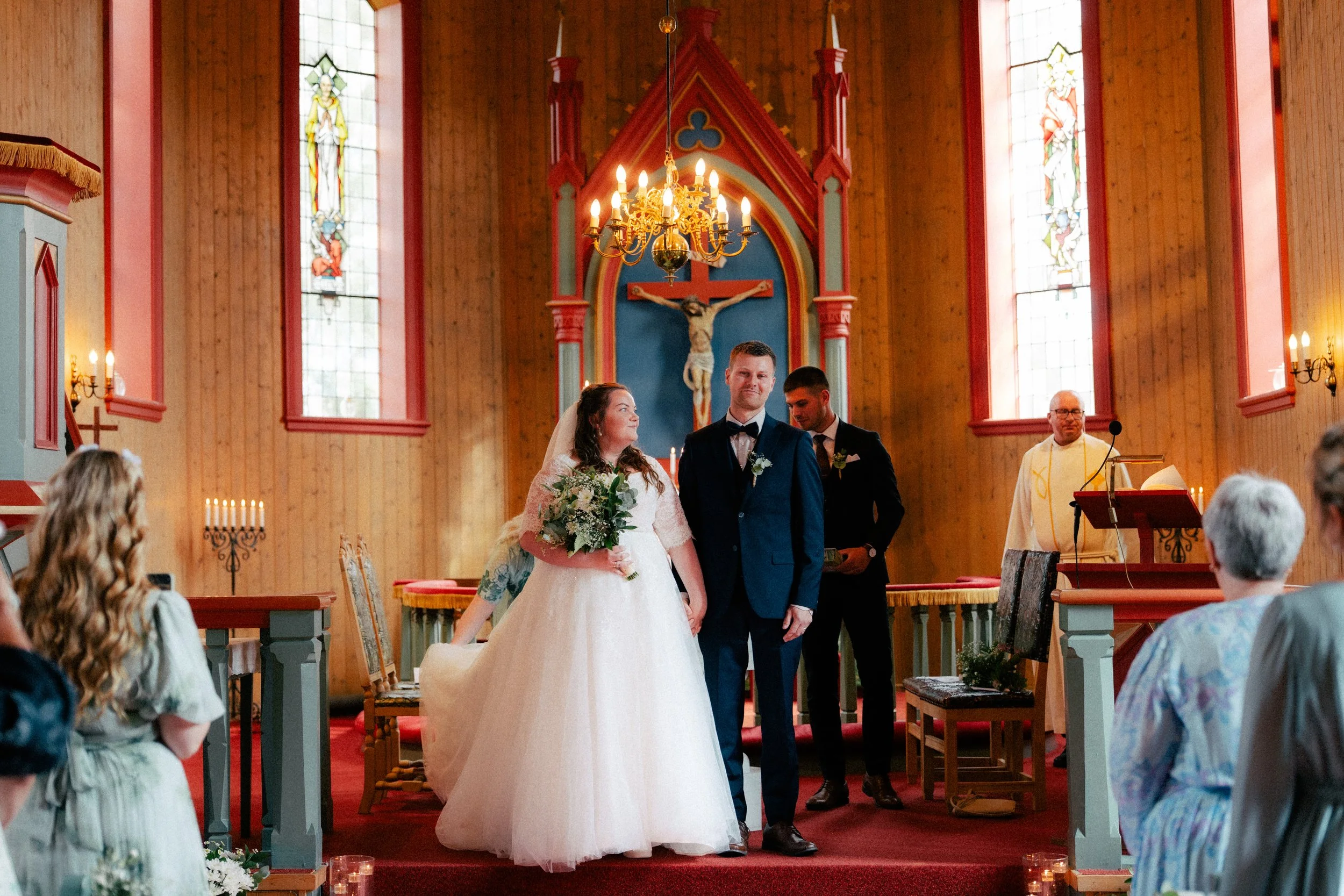 En vielsesser som står ved alteret i en kirke, omgitt av venner og familie, under en seremoni. Kvinnen bærer en hvit brudekjole og holder en blomsterbukett, mens mannen ved siden av henne har på seg en mørk dress og slips. Bakgrunnen viser en kors me