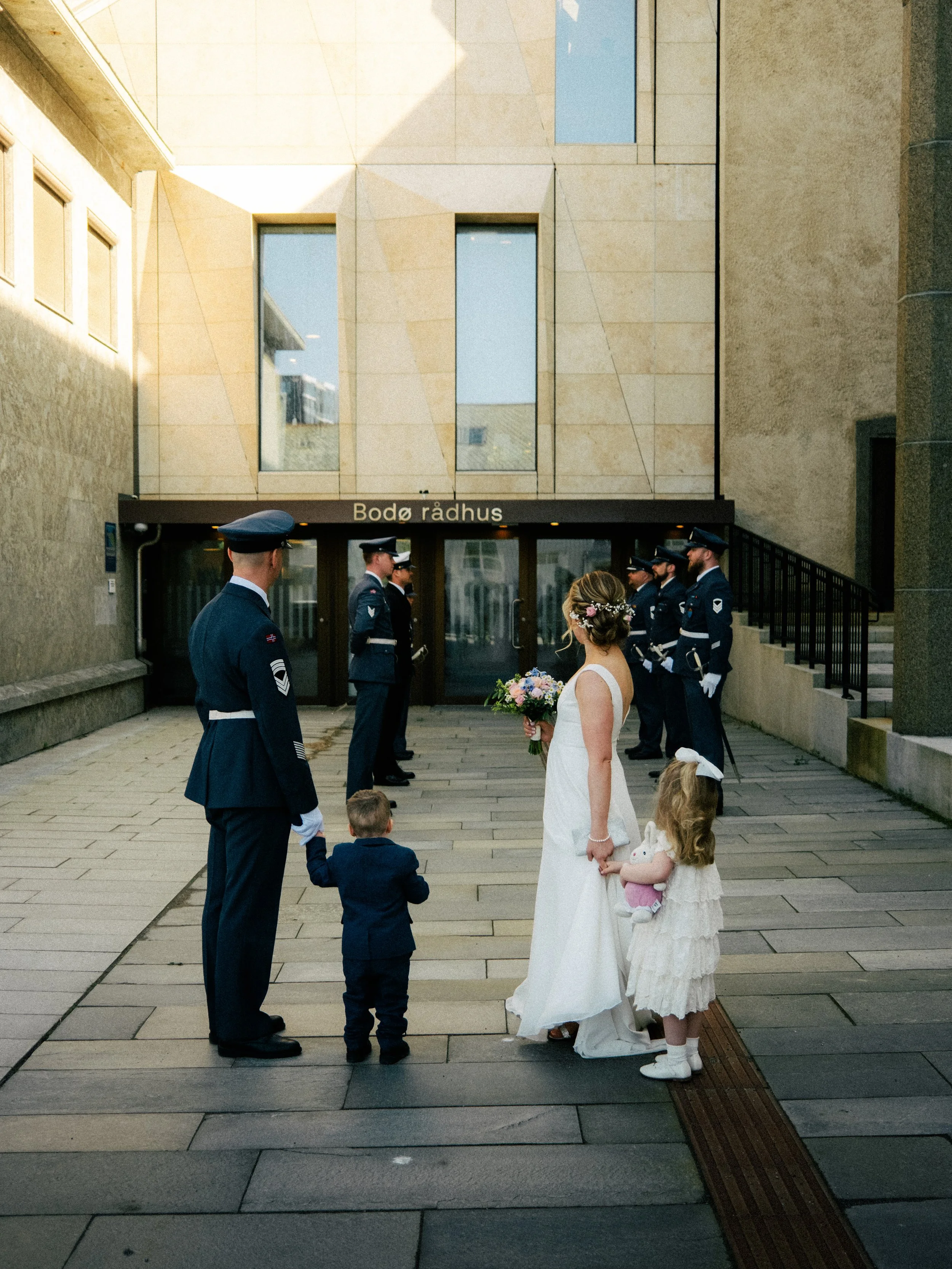 En gruppe mennesker, inkludert to barn, står foran rådhuset. En dame i hvit kjole holder en blomsterbukett og holder hendene til en liten jente i hvit kjole. Flere menn i militæruniformer står i bakgrunnen.