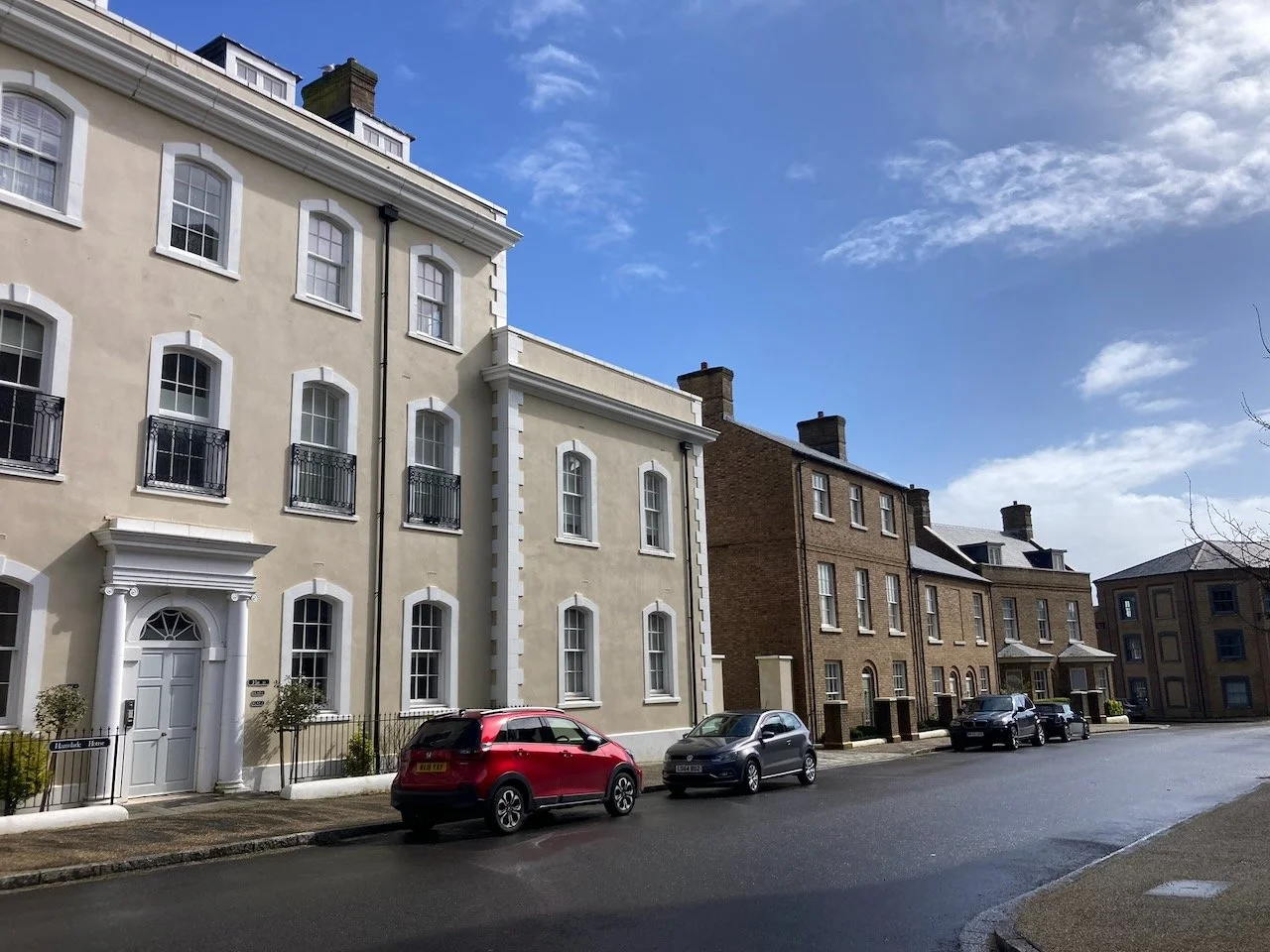 Residential street with modern cream-colored and brown brick townhouses, parked cars, and partly cloudy sky.