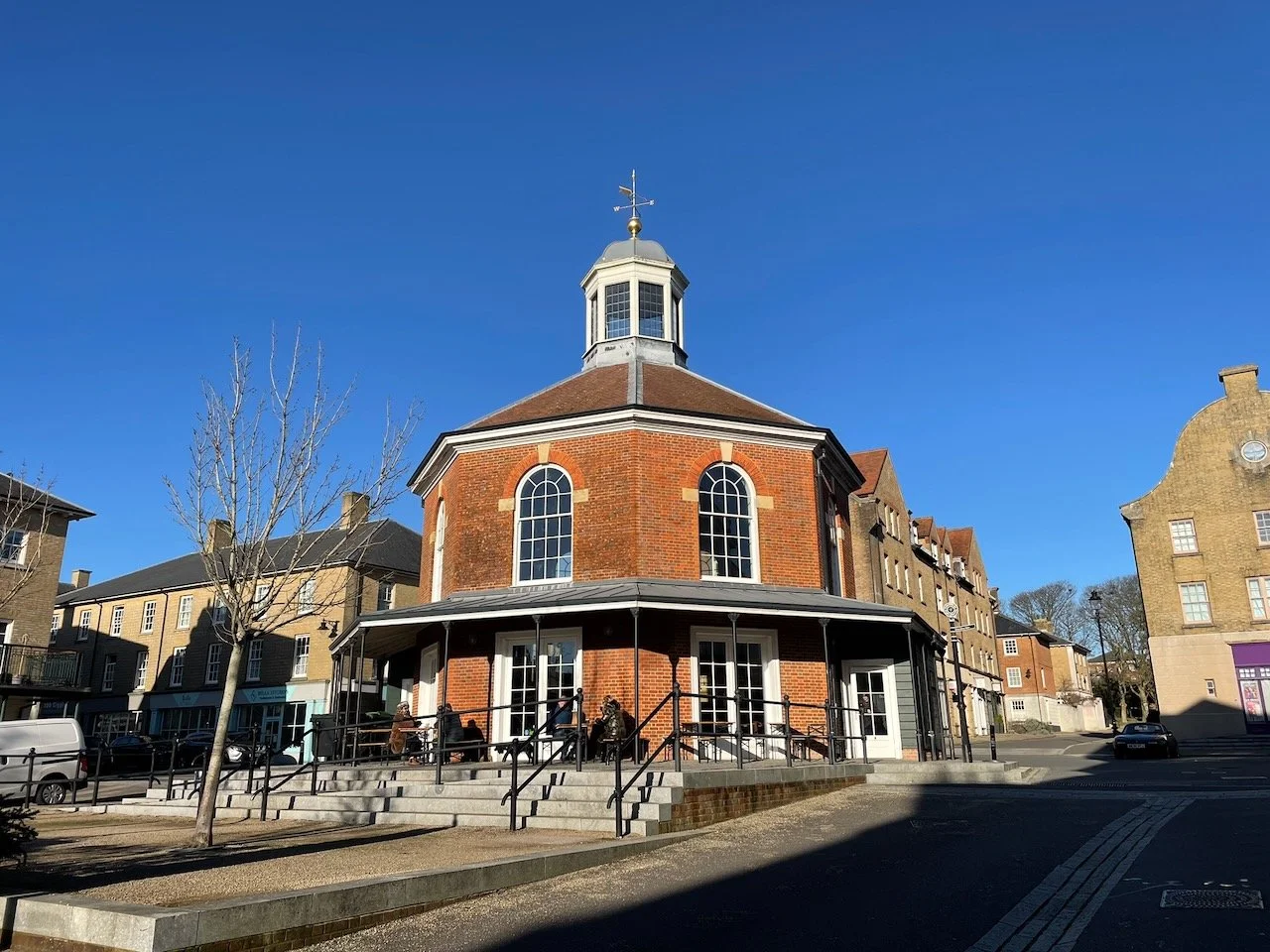 A red brick building with a round shape and tall arched windows, topped with a small cupola and weather vane, surrounded by a small outdoor seating area with people, in a town square with other buildings and cars.