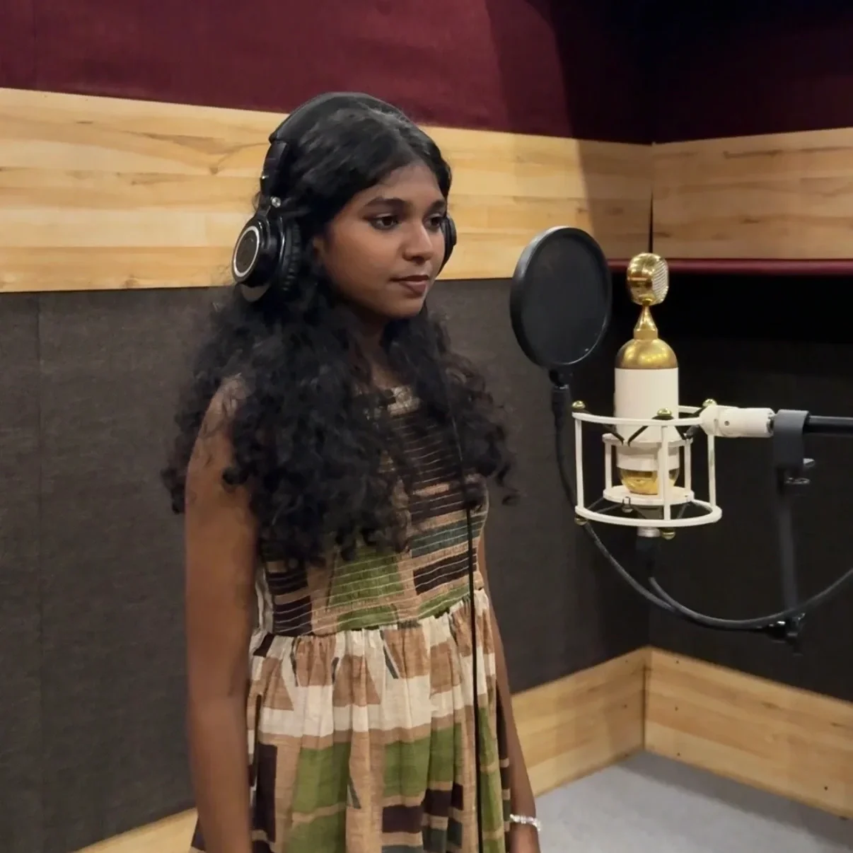 A woman with long, dark curly hair wearing headphones, standing in a recording studio in front of a microphone with a pop filter.