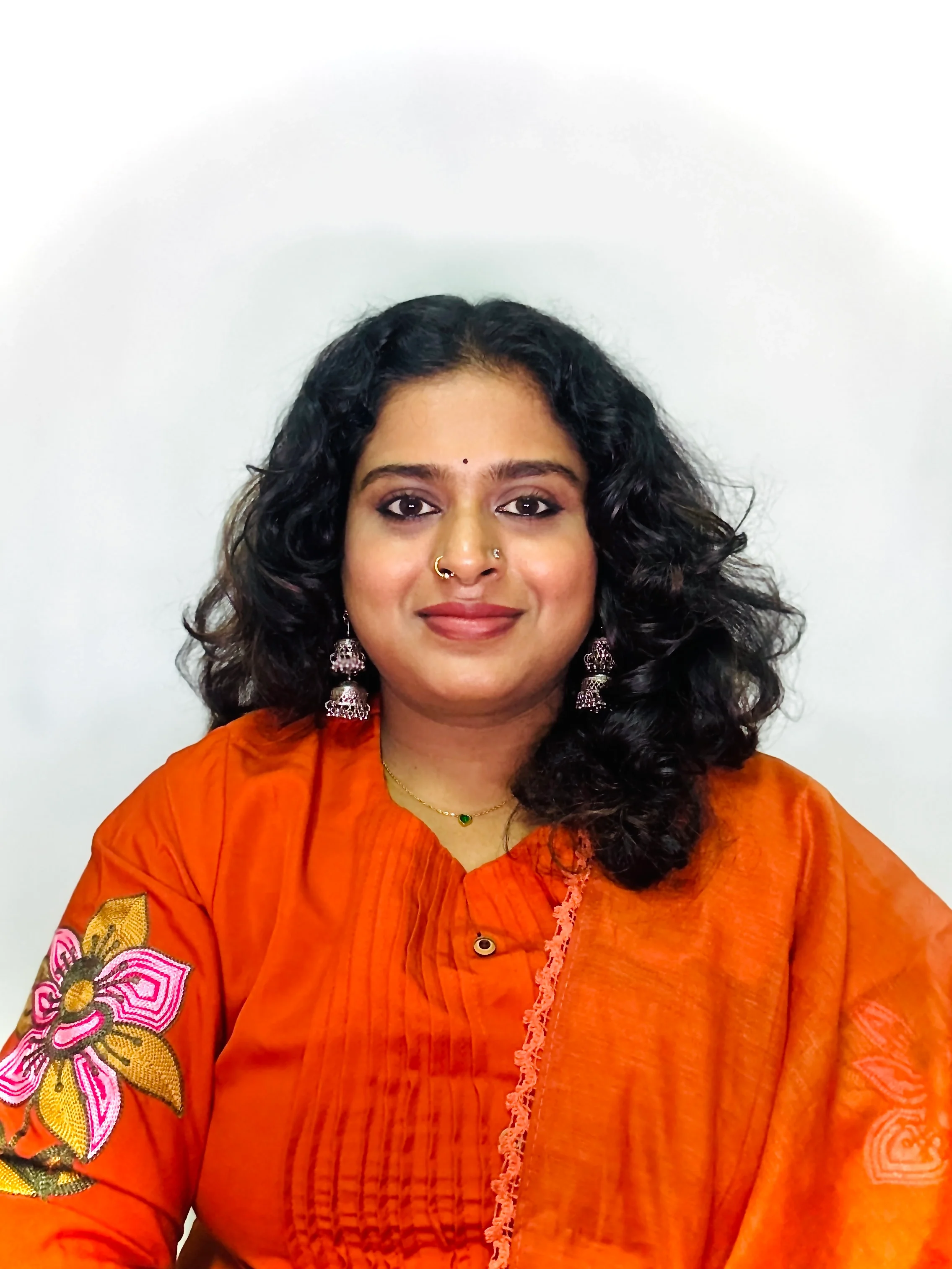 A woman with curly black hair, wearing a bright orange traditional Indian outfit with floral embroidery, jewelry including earrings, a nose ring, a necklace, and a small bindi on her forehead, smiling against a plain white background.