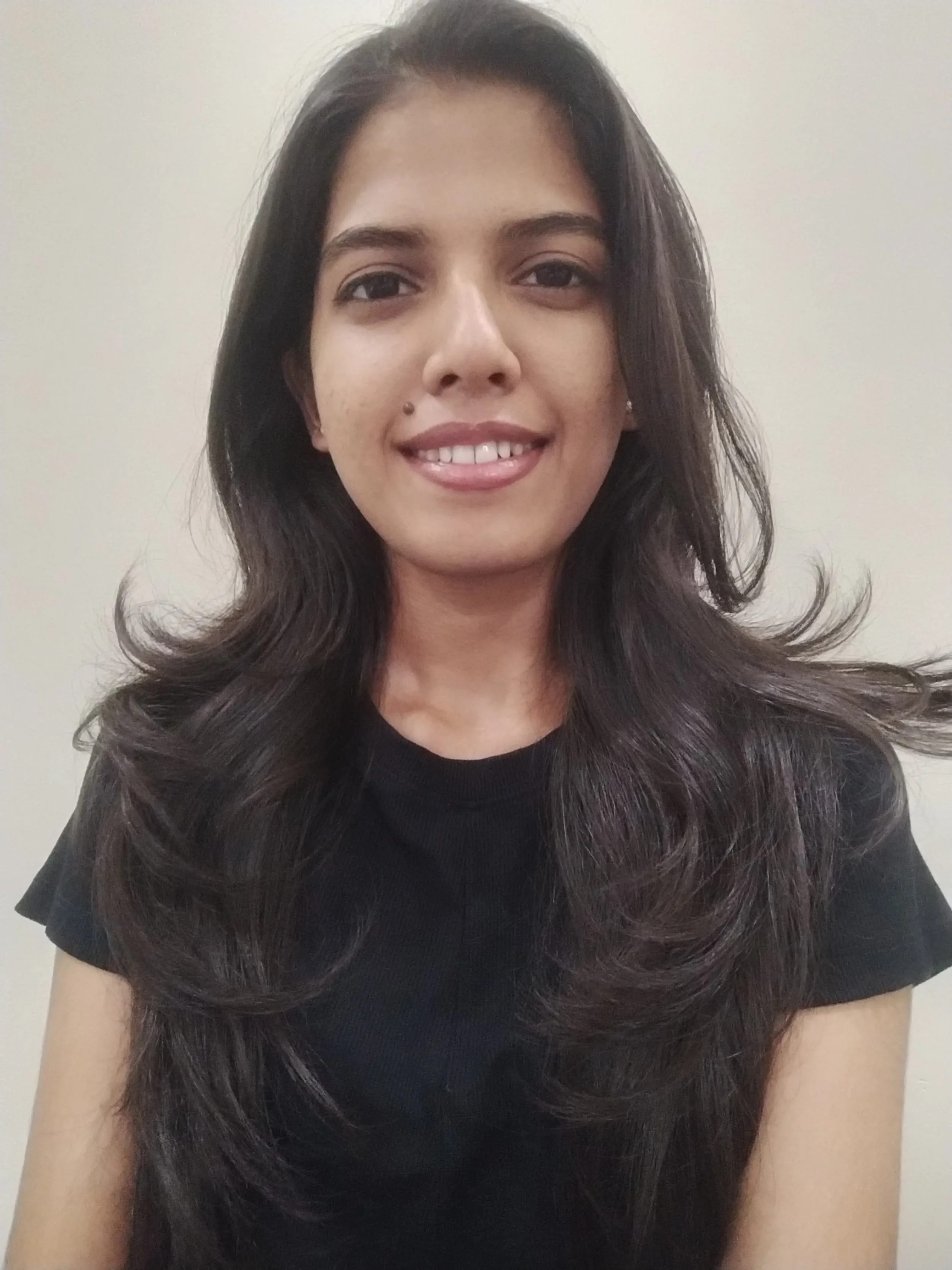 A young woman with long, dark, wavy hair, smiling at the camera against a plain, light-colored background.