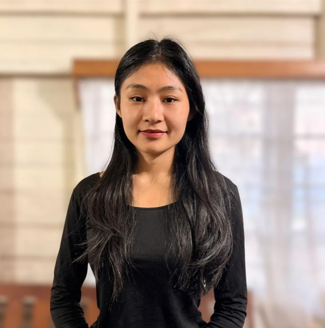 A young woman with long black hair wearing a black top standing indoors against a blurred background.