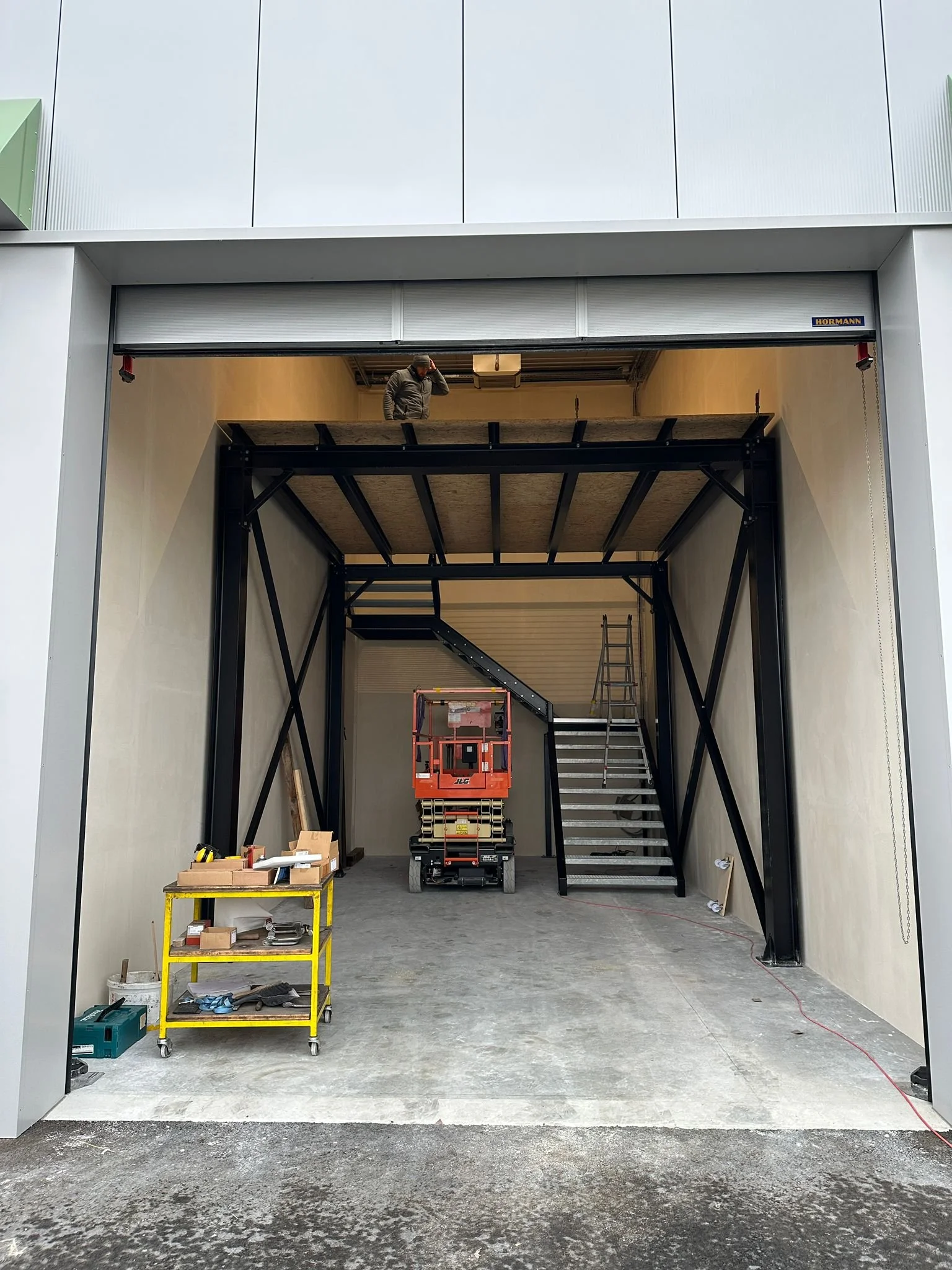 Der Innenraum eines Garagentors, mit Bauarbeitern, einer Leiter, einem Rolllift und Werkzeugen, die an verschiedenen Stellen im Raum platziert sind.