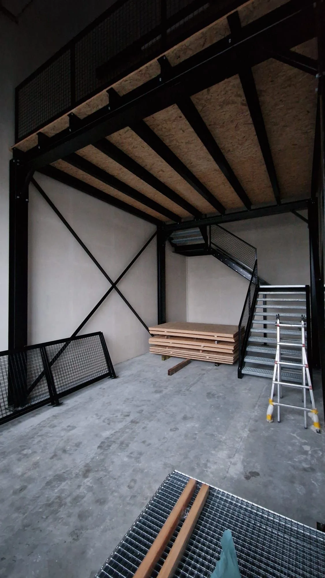 Ein unzureichend fertiggestelltes Hochbett in einem Raum, mit Holzplatten, einer Leiter, Staubbrettern und Metallgittern.