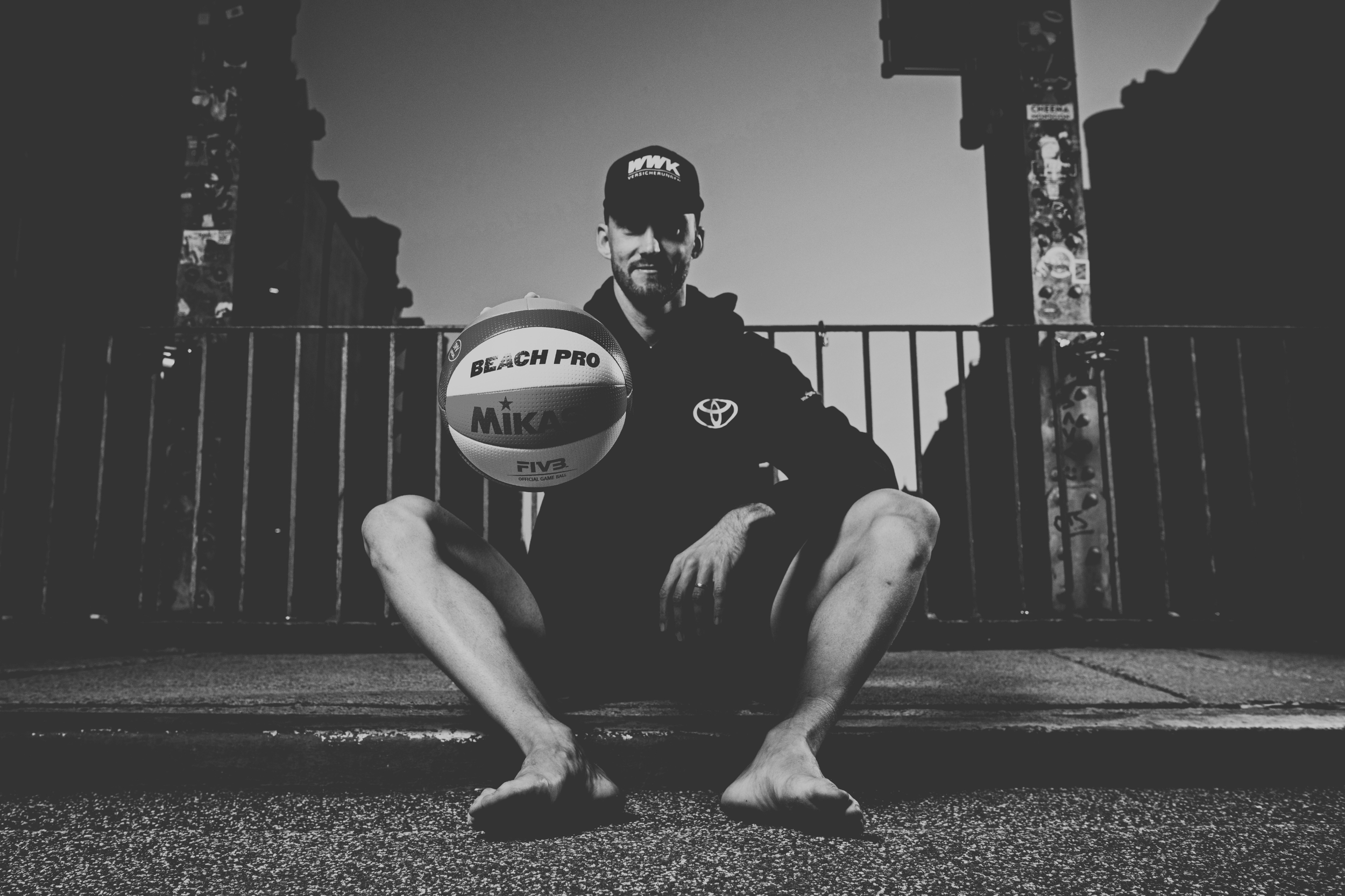 Ein Mann sitzt barfuß auf dem Boden, hält einen Volleyball vor sich und blickt lächelnd in die Kamera. Er trägt eine schwarze Kappe und eine schwarze Jacke mit Toyota-Logo, im Hintergrund eine Metallabsperrung und eine Brücke.