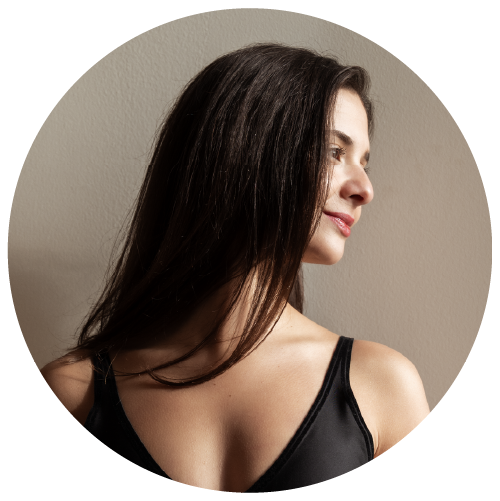 A woman with long dark hair looking to the right, wearing a black sleeveless top, against a plain beige wall.