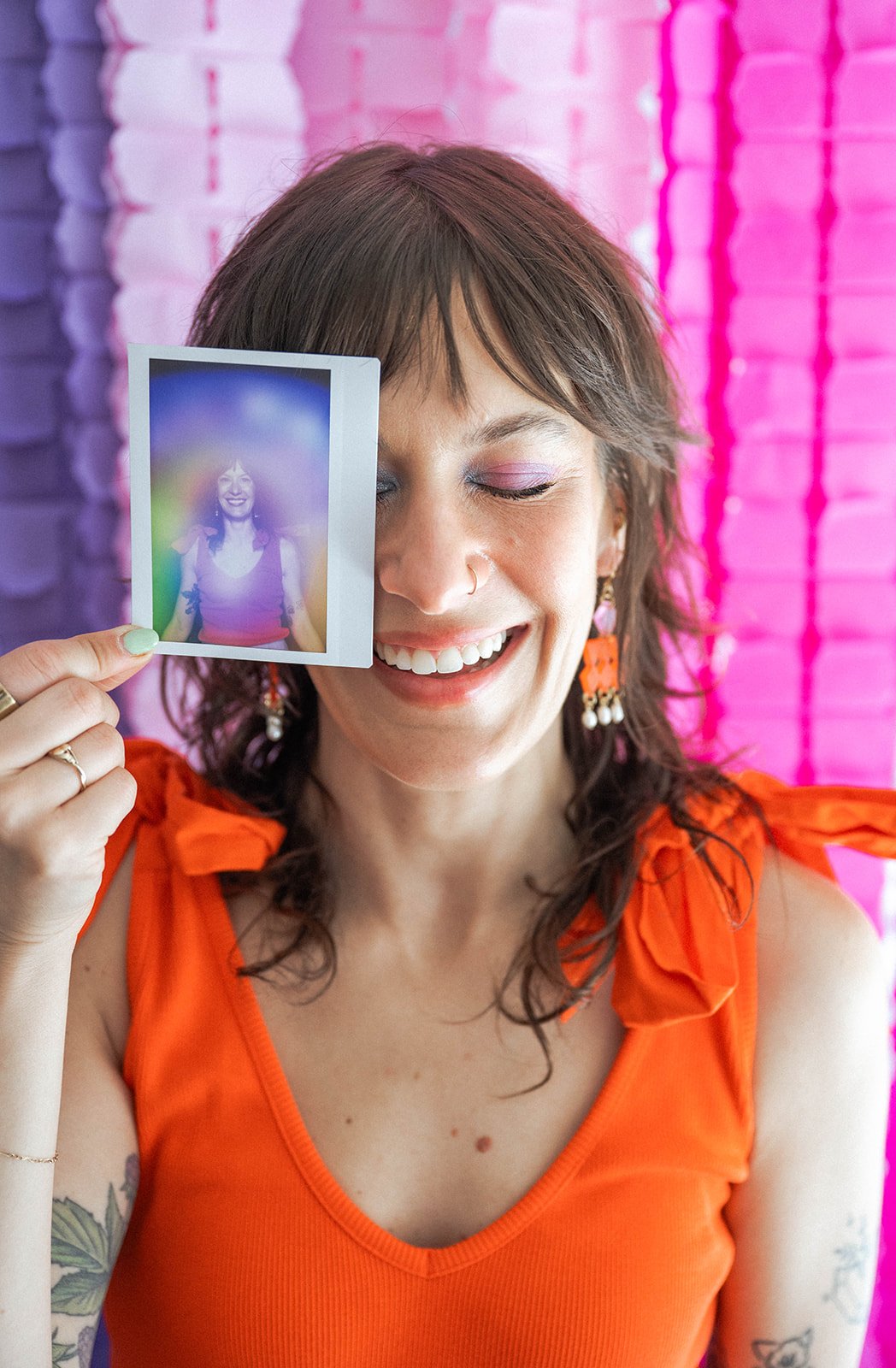 A woman with short brown hair and colorful earrings smiling with her eyes closed, holding a Polaroid photo in front of her face. The background is a pink brick wall.