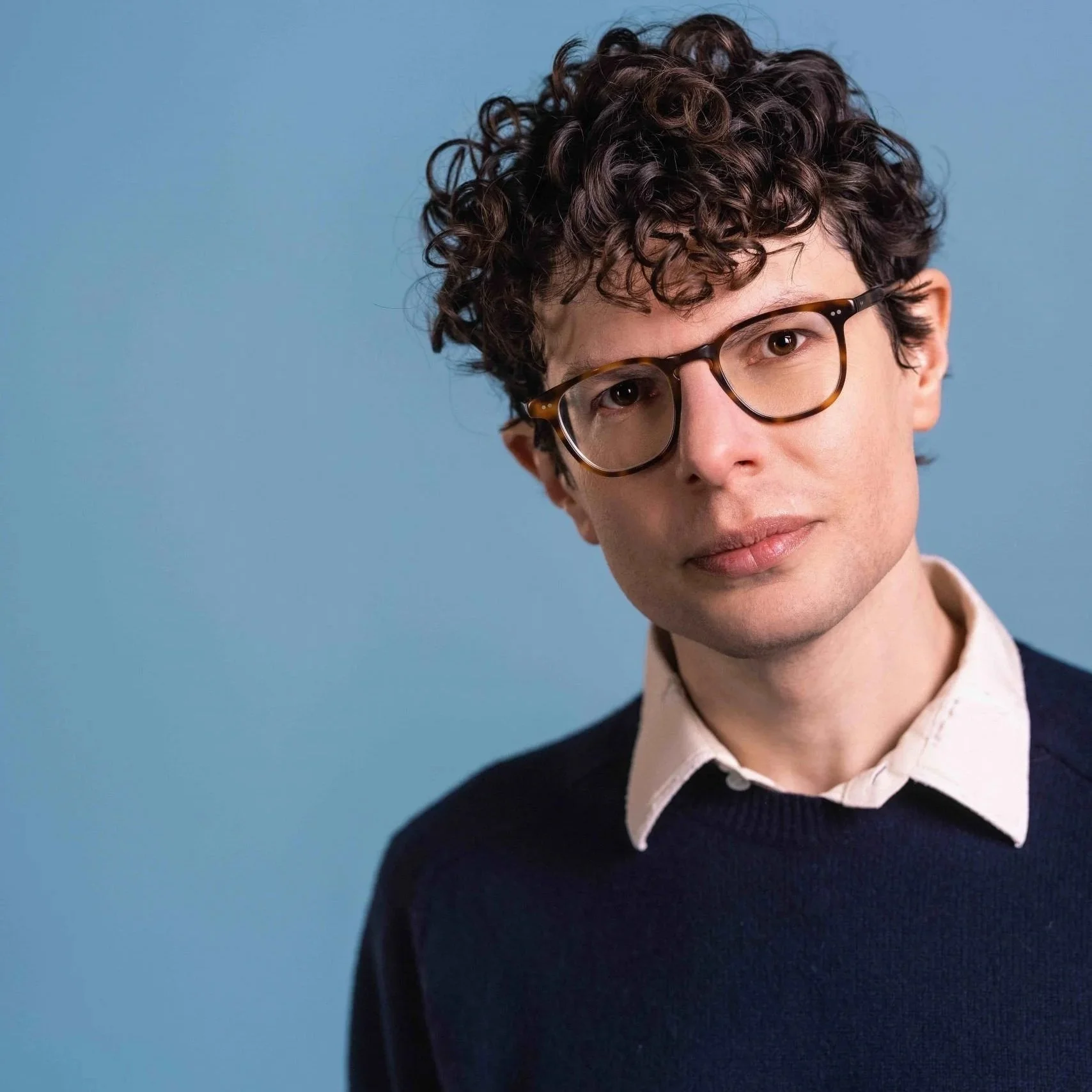 Portrait of a young man with curly brown hair and glasses, wearing a cream collared shirt under a dark sweater, against a blue background.