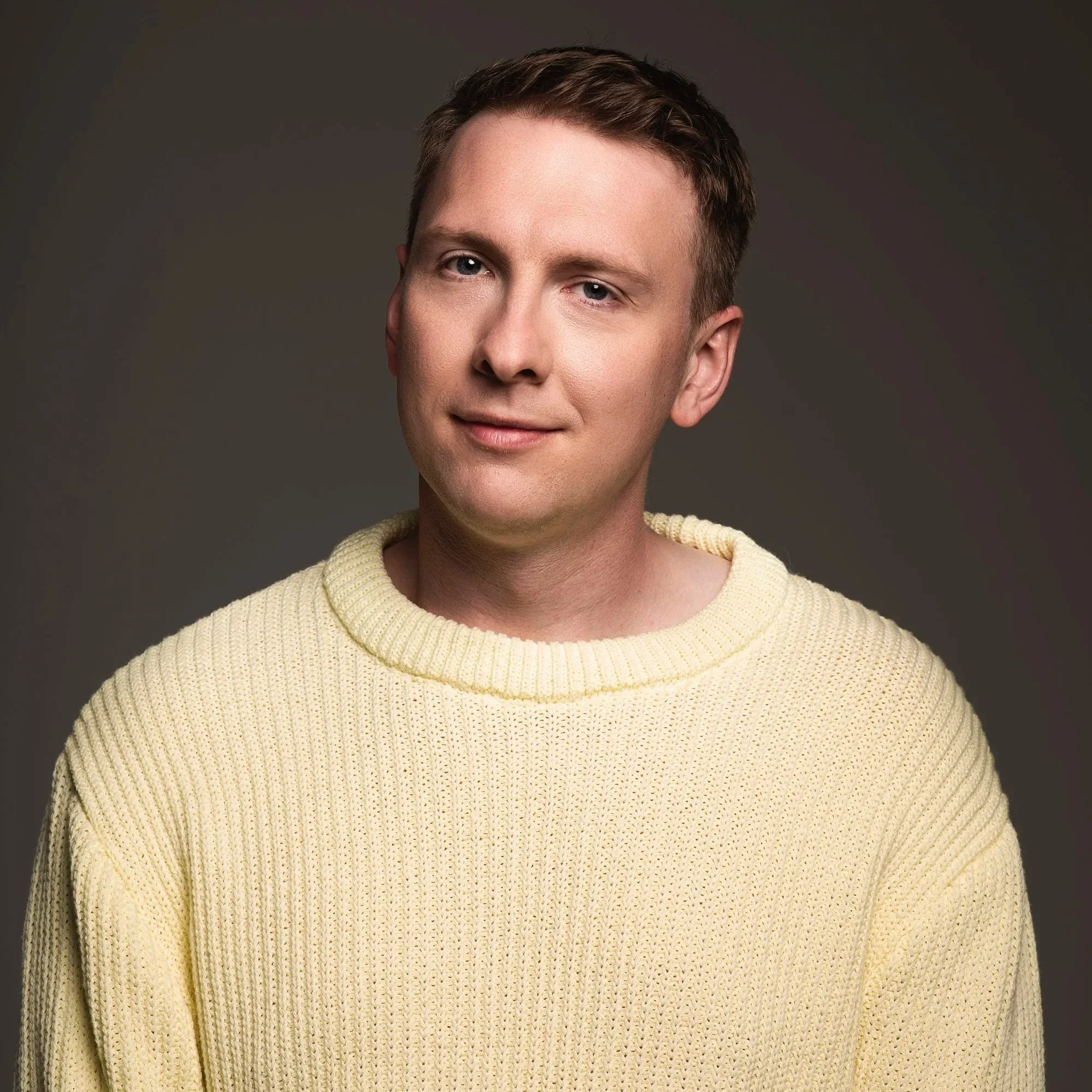 Portrait of a young man with short brown hair, blue eyes, wearing a cream-colored knit sweater, smiling gently against a dark background.