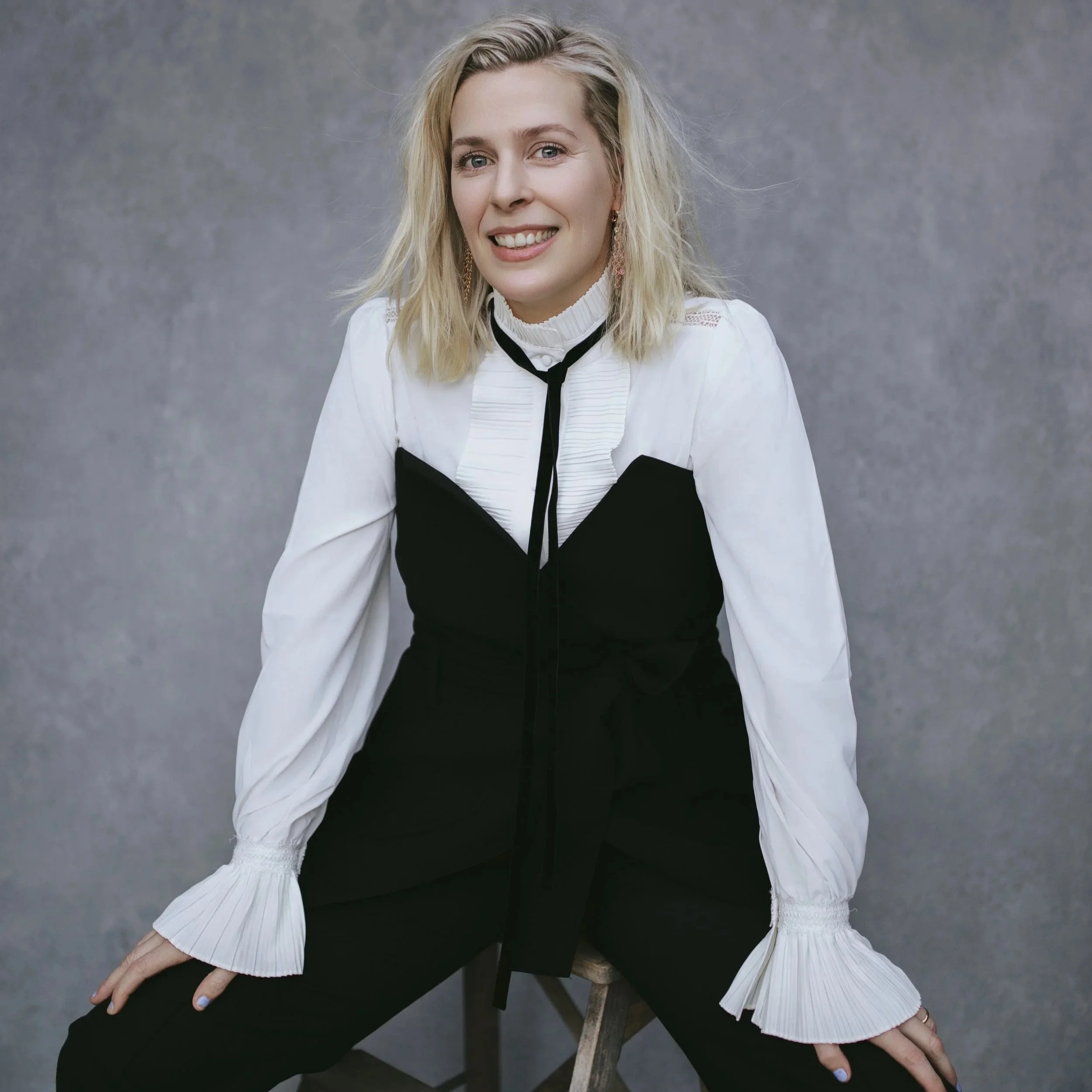 A woman with blonde hair smiling, sitting on a stool, wearing a black and white outfit against a gray textured background.