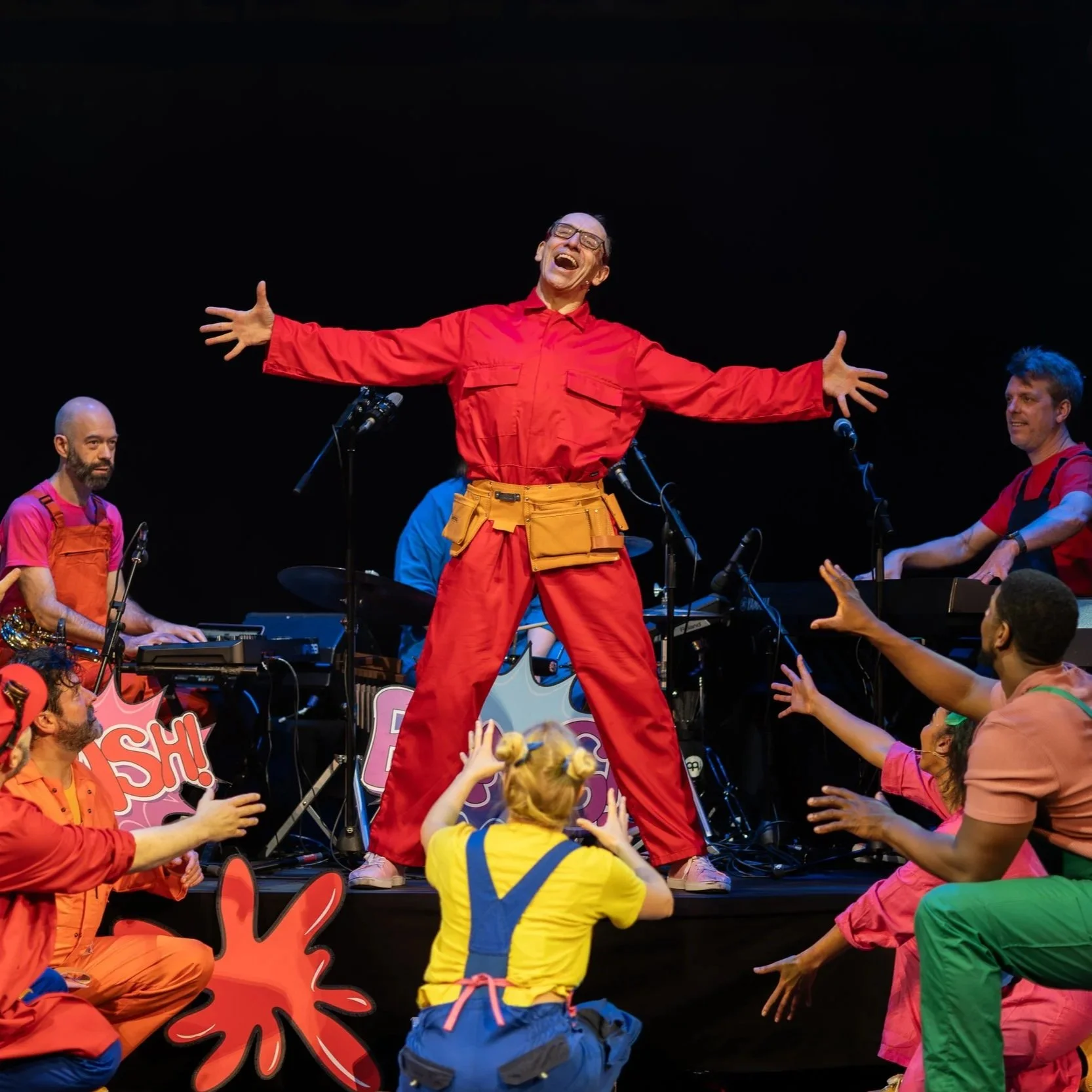 Performer on stage singing with arms outstretched, surrounded by colorfully dressed audience members reaching towards them, with musicians playing instruments in the background.