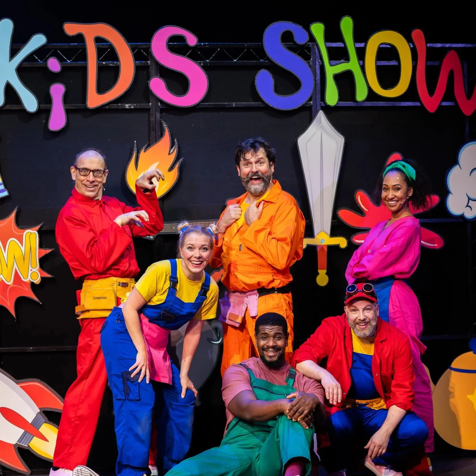 Group of six people dressed in colorful costumes posing in front of a backdrop with children's show related decorations and text 'Kids Show'.