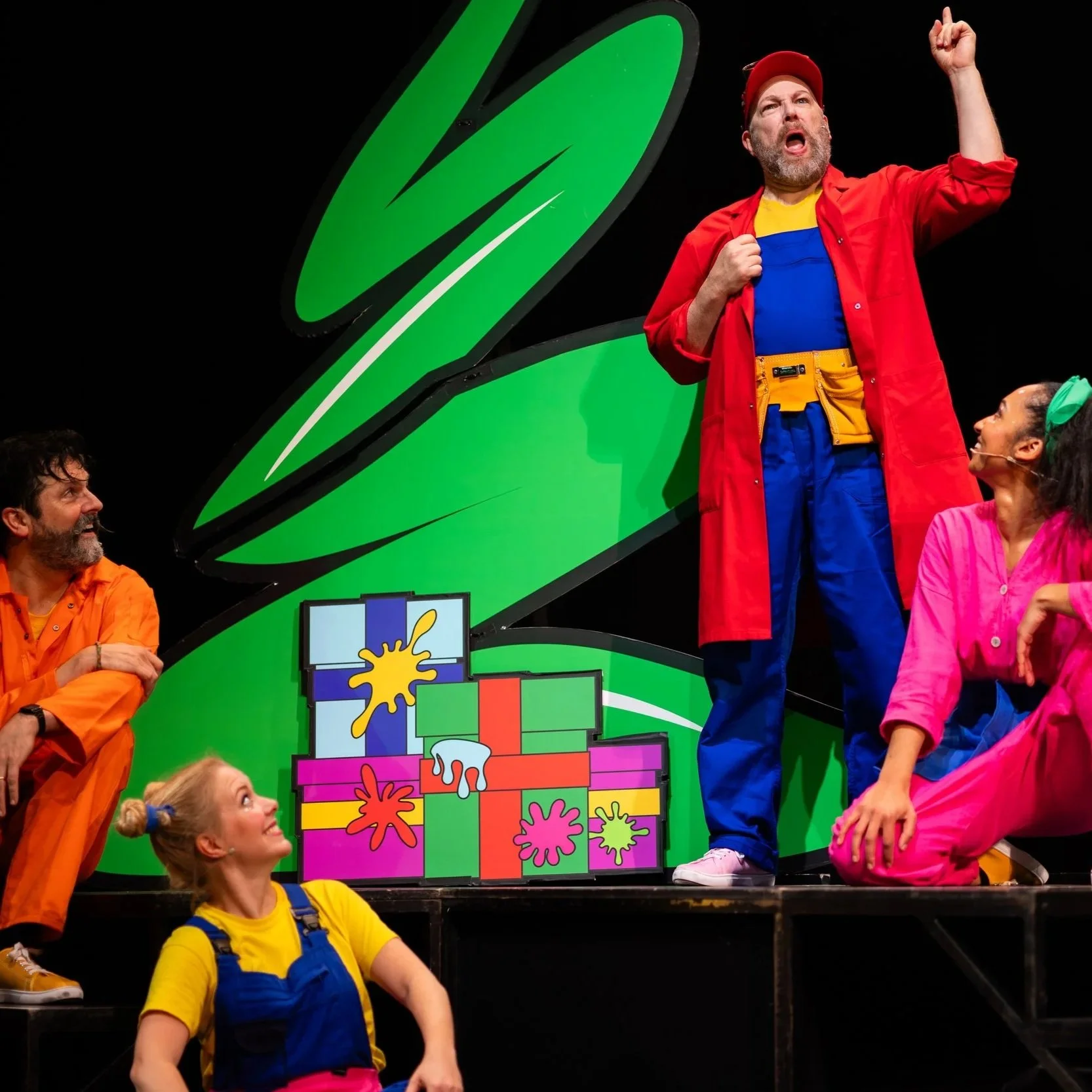 Actors performing a colorful stage play with large greenery and painted gift boxes, some actors dressed in bright costumes.